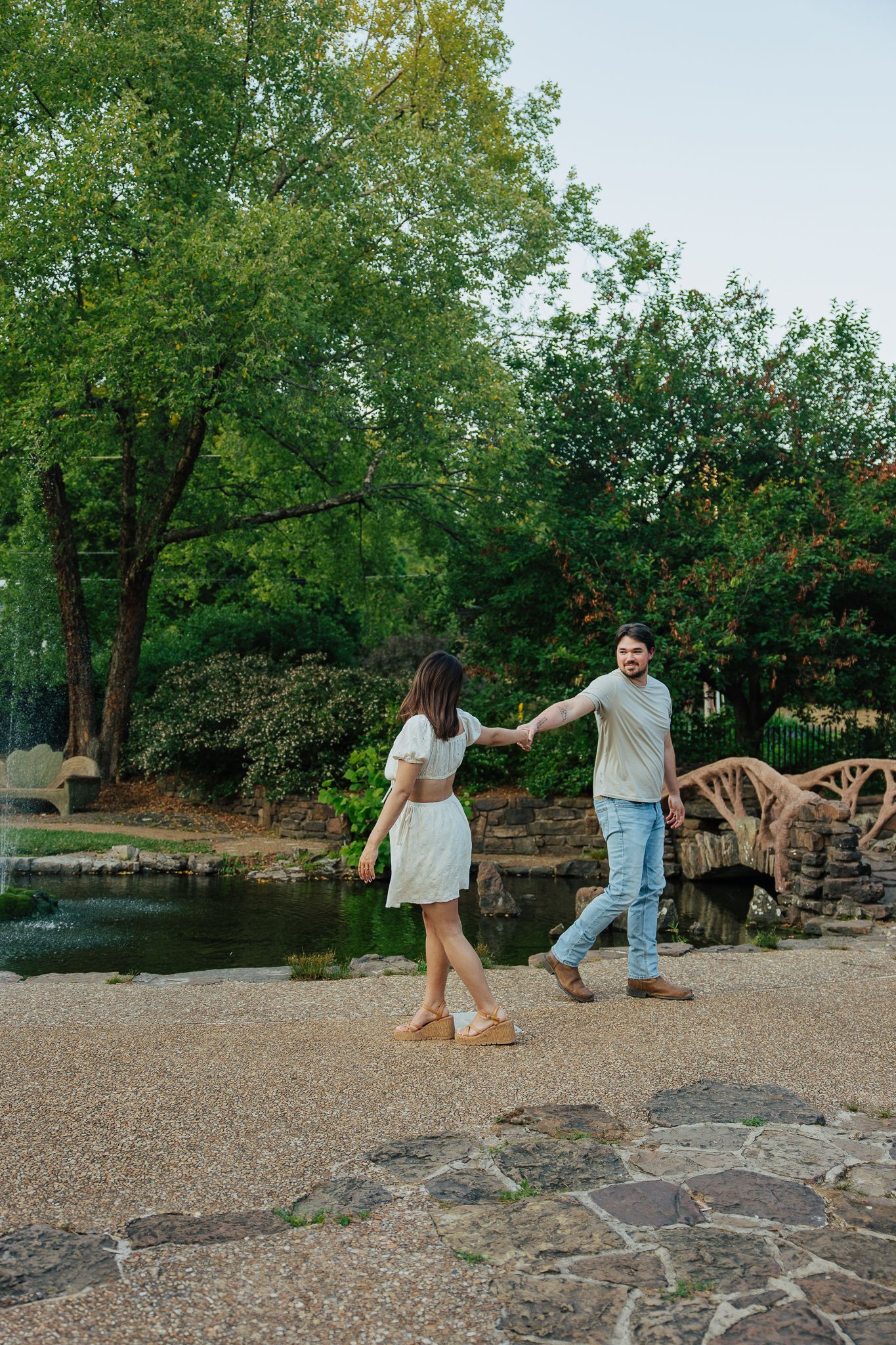 A couple holds hands walking through a lush green park near a decorative stone bridge.