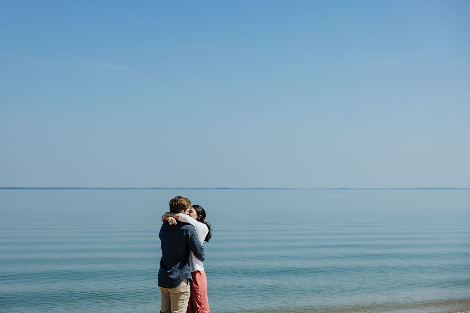 Couple embracing on serene beach shore under clear blue sky with calm ocean behind.