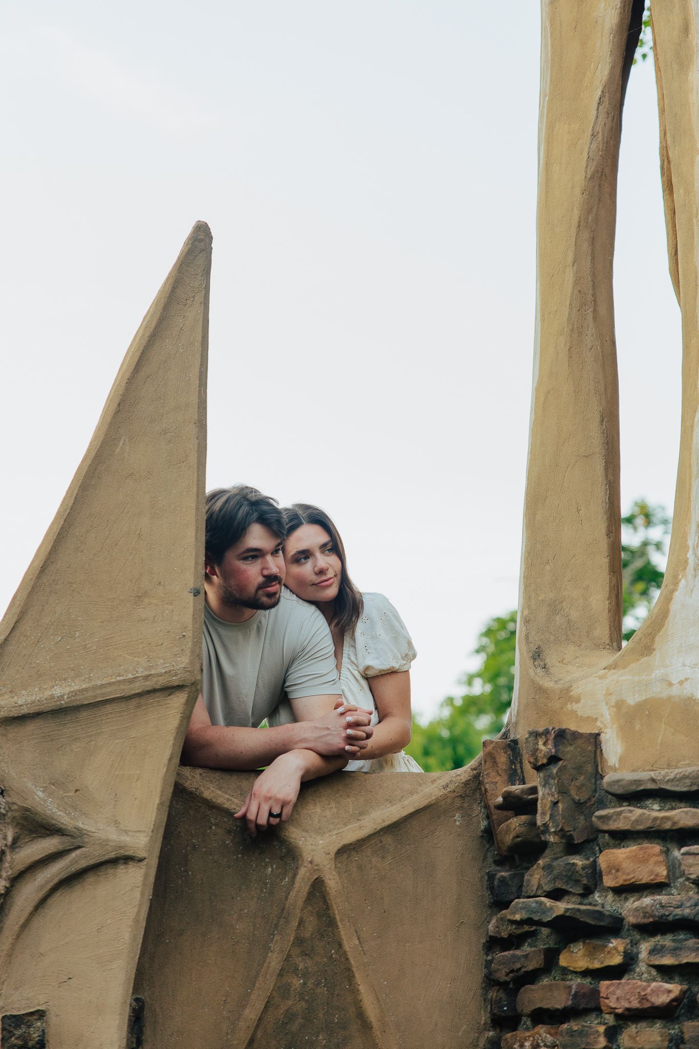 Couple embracing and leaning against angular stone sculptures while gazing outward in garden park.