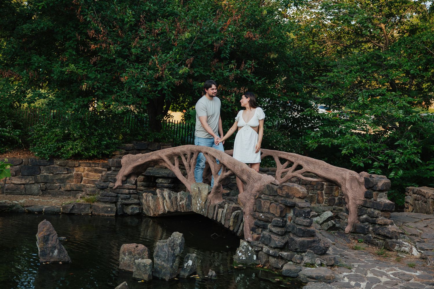Couple holds hands on rustic stone bridge over pond surrounded by lush green trees during engagement session.