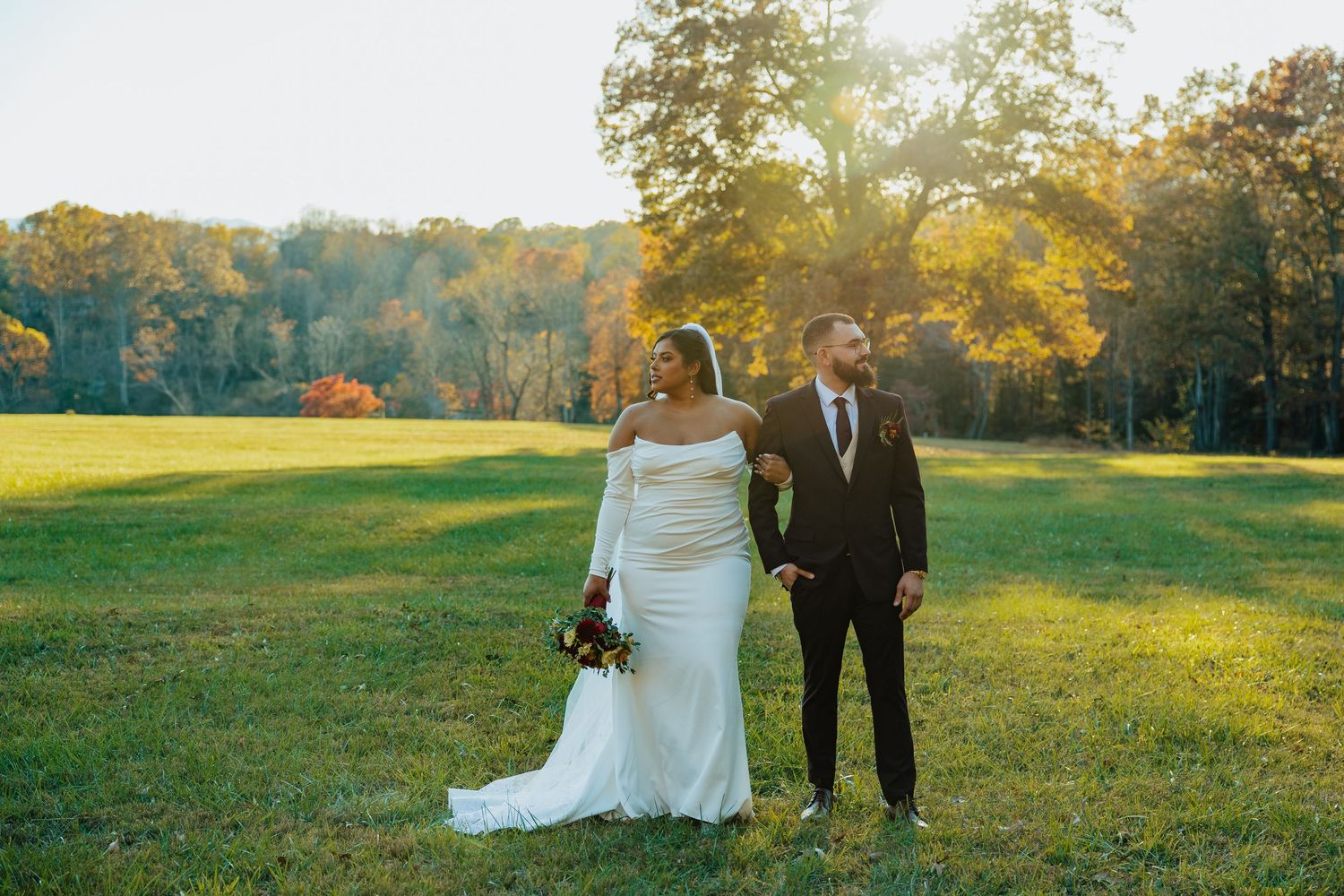 Bride and groom pose together in a golden sunlit meadow surrounded by colorful fall foliage.