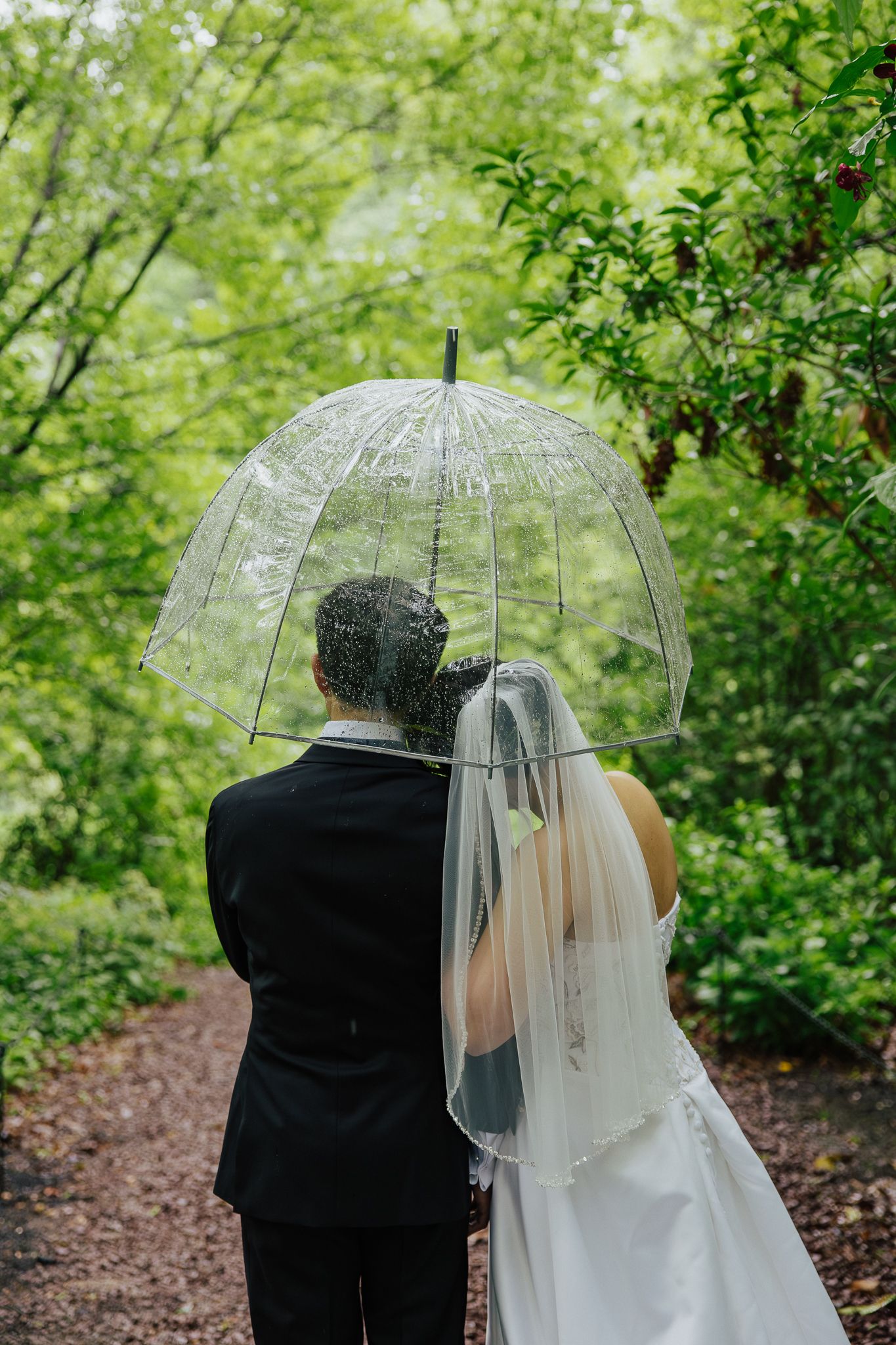 A bride and groom share an umbrella walking through a lush green forest path on a rainy wedding day.
