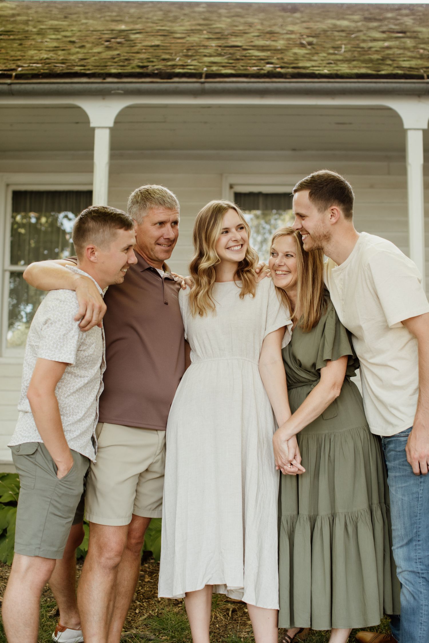 Five family members smiling and embracing outdoors in front of a white porch home during a casual family portrait session.