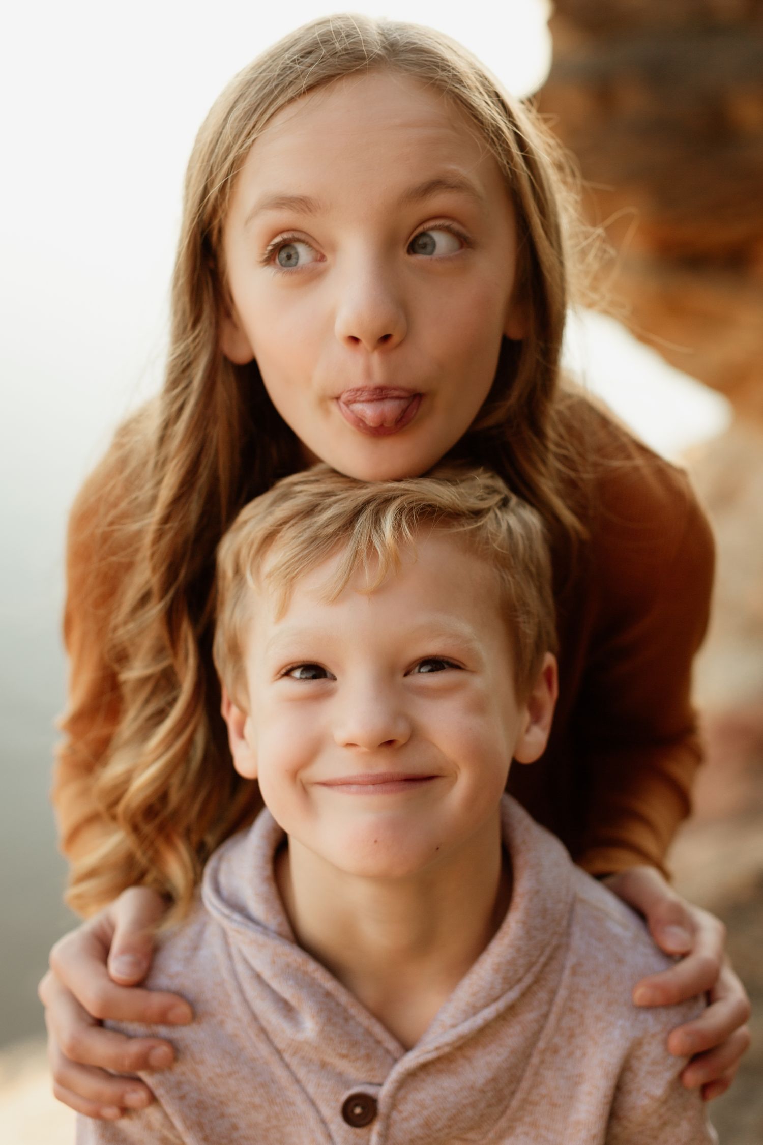A teenage girl sticking out her tongue hugs a smiling young boy from behind in a playful outdoor portrait.