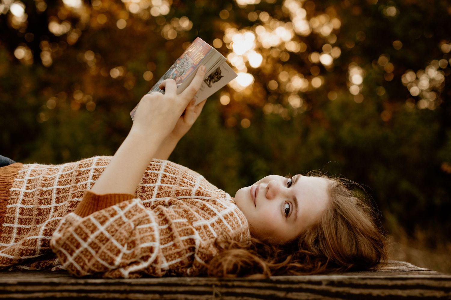 Young woman lying on wooden surface reading a book in warm golden bokeh light.