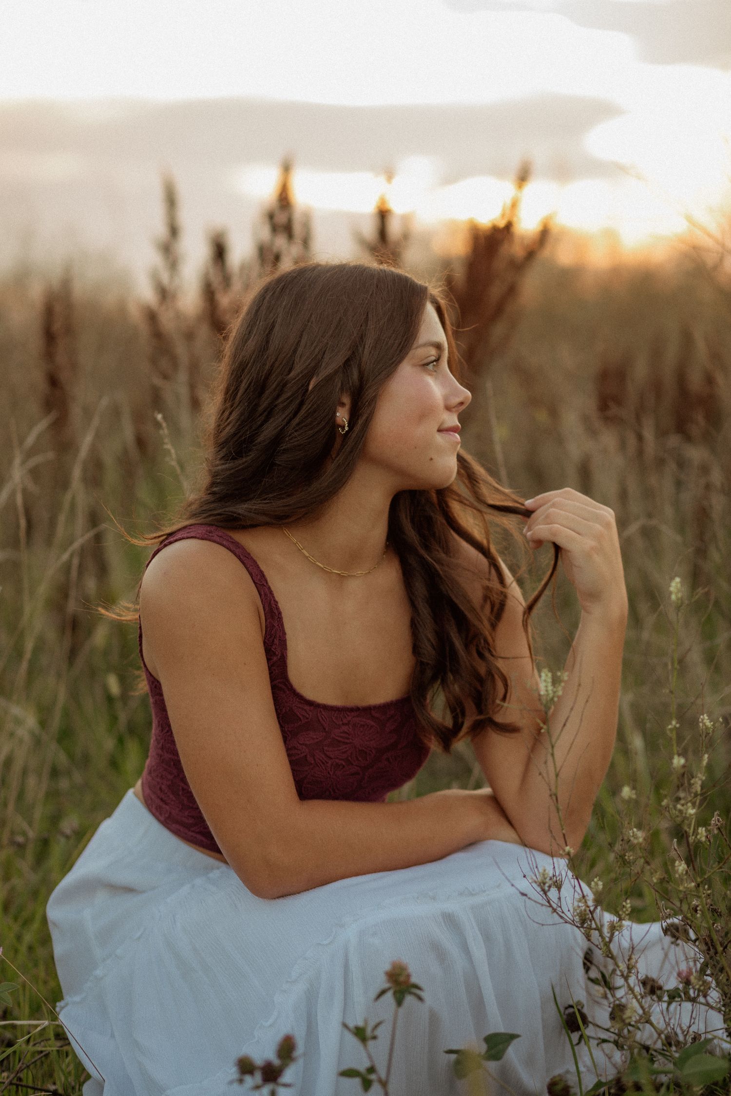 Brunette woman in burgundy top and white skirt sitting in tall grass field at golden sunset.