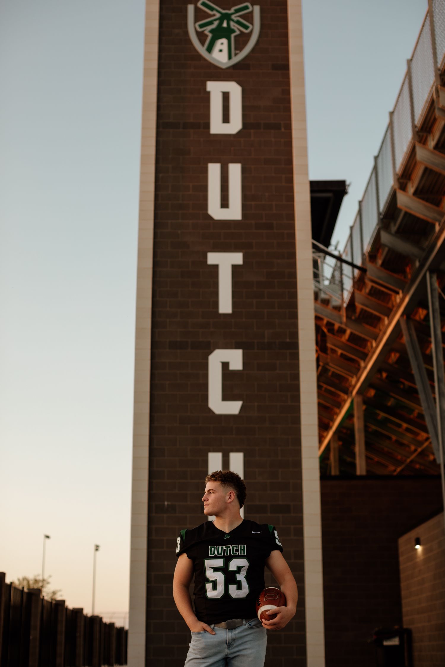 Young man in Dutch #53 football jersey standing beside a tall brick DUTCH stadium tower at dusk.