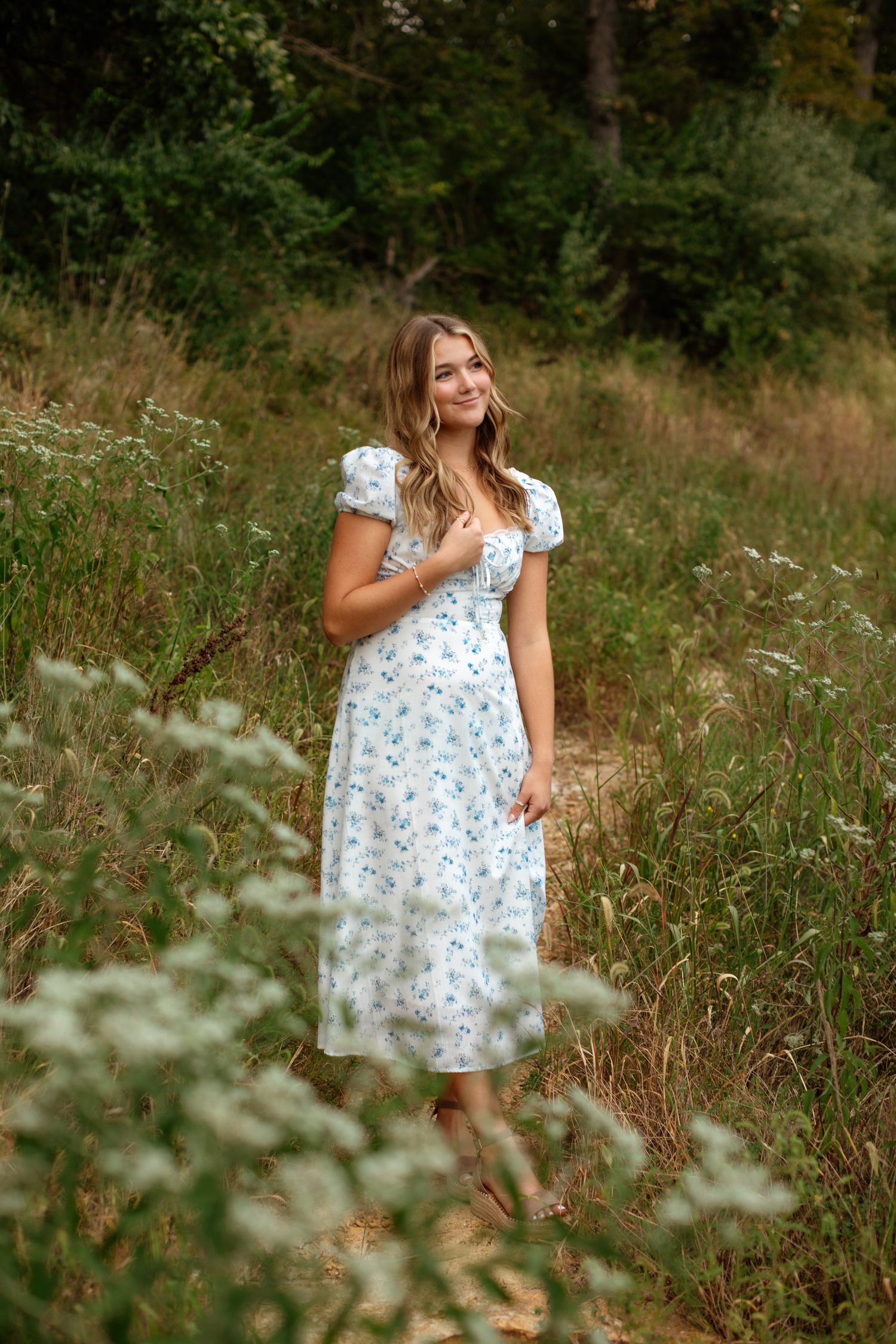 Young woman in white floral midi dress standing in a wildflower meadow surrounded by tall grass and trees.