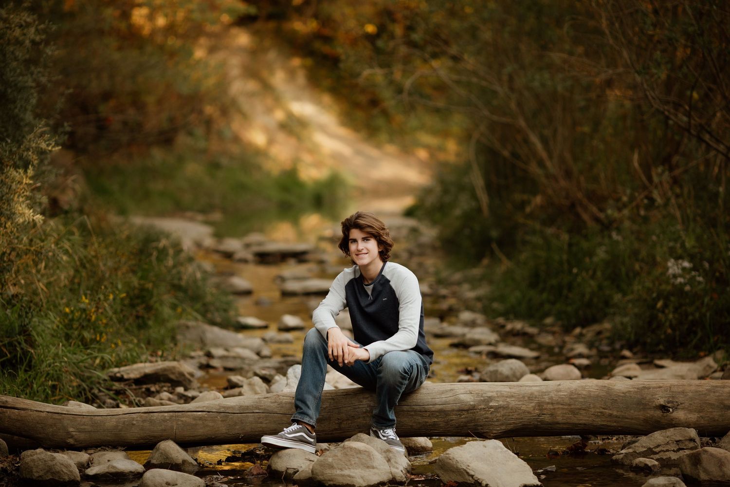 Teenage boy with curly hair sitting on a log by a rocky creek in an autumn forest.