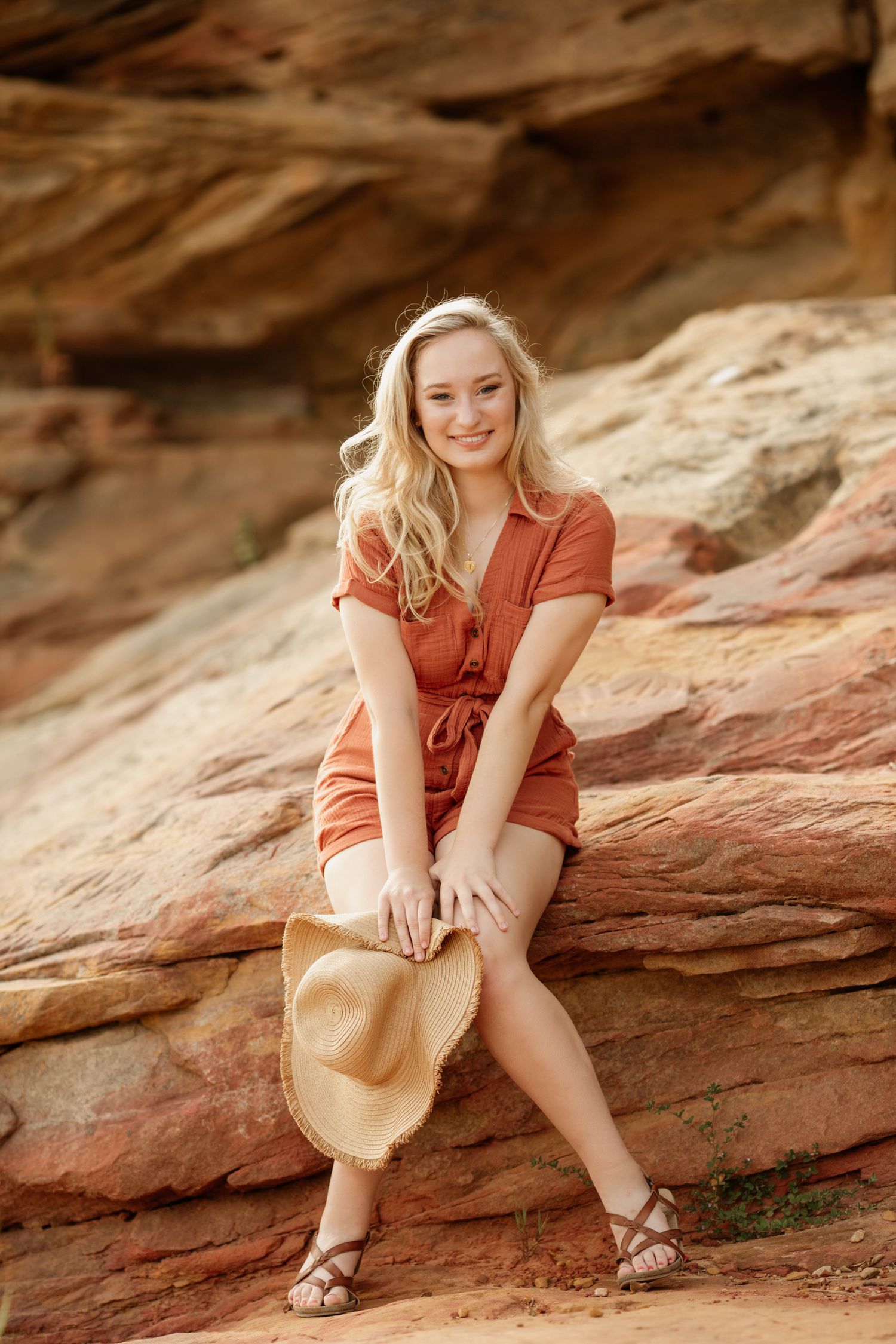 Smiling blonde teen in rust orange romper sits on red rock formations holding a straw hat.