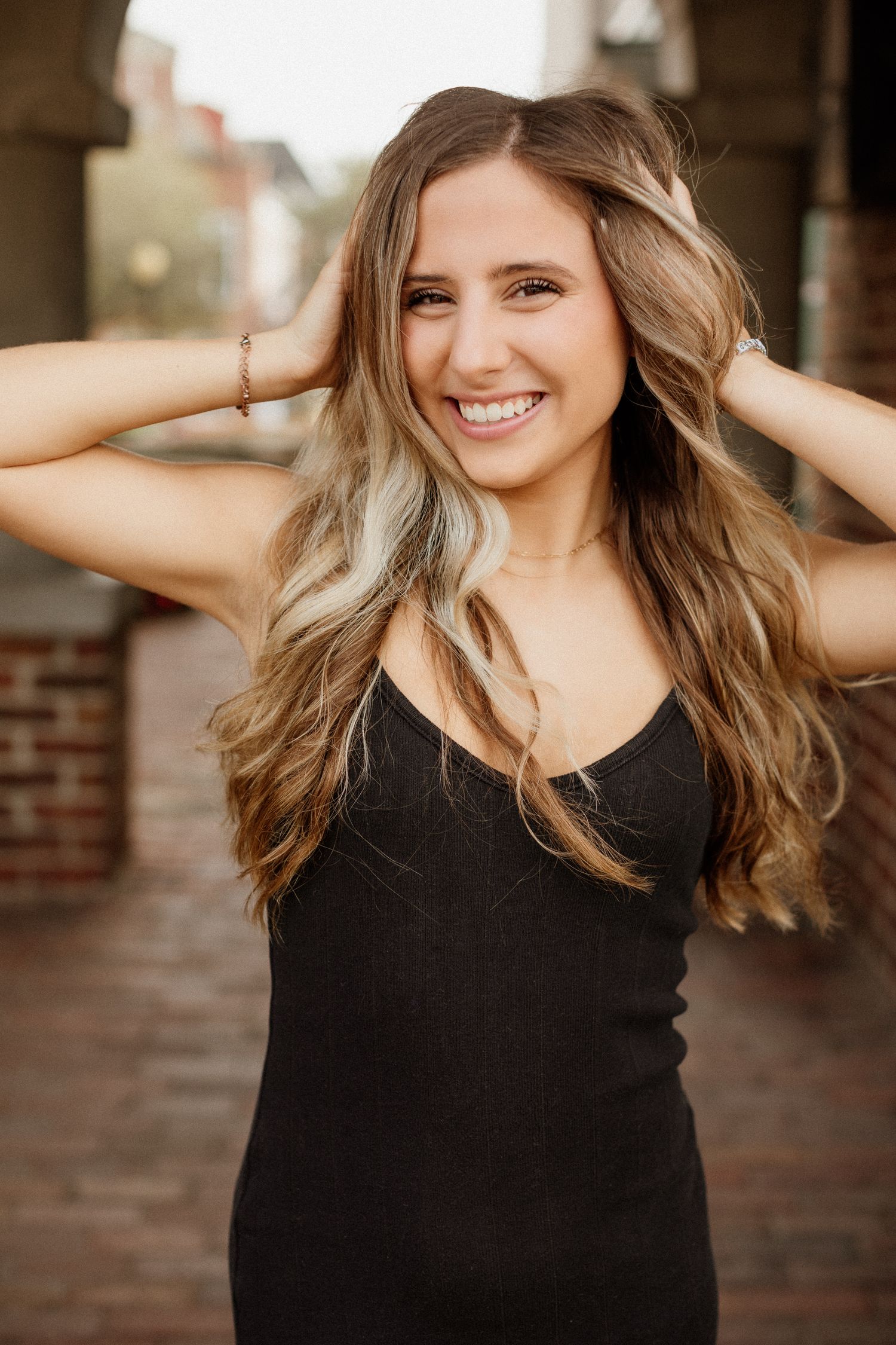 Smiling young woman with long wavy hair in black dress running hands through hair on a brick walkway.