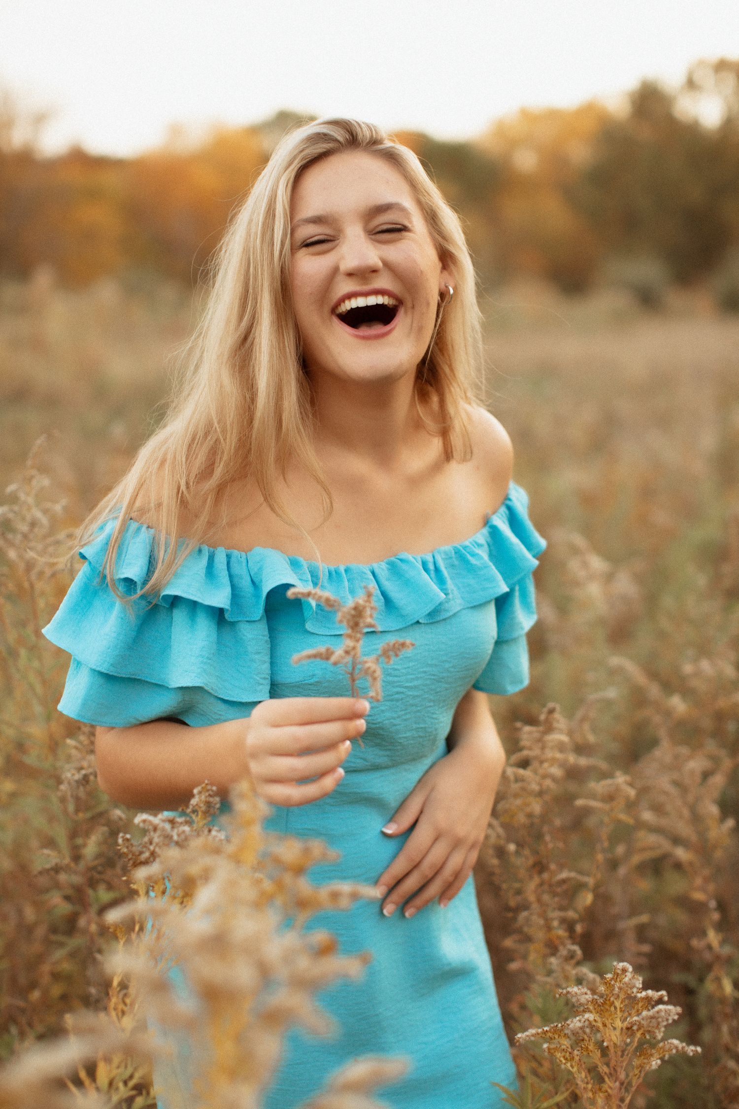 Joyful blonde woman in a blue off-shoulder dress laughing in a golden autumn field.