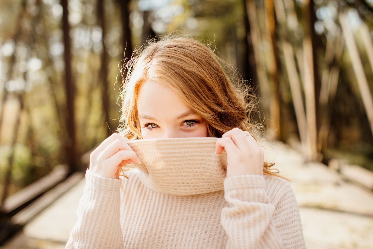 Young woman with auburn hair pulls a cream turtleneck sweater over her face in a sunny forest.