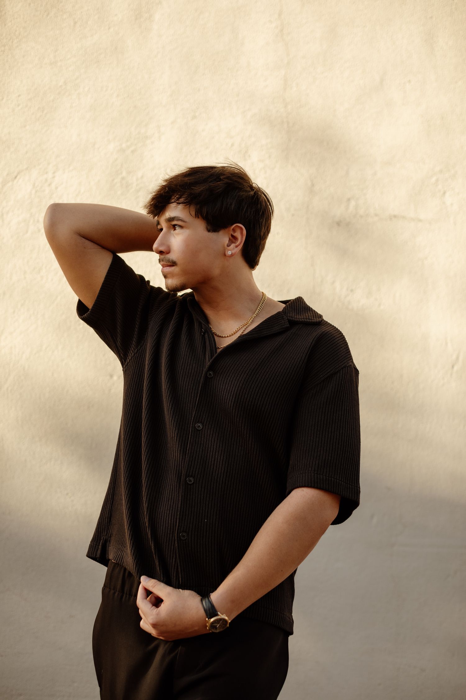 Young man in black short-sleeve button shirt and watch posing against a warm-lit textured beige wall.