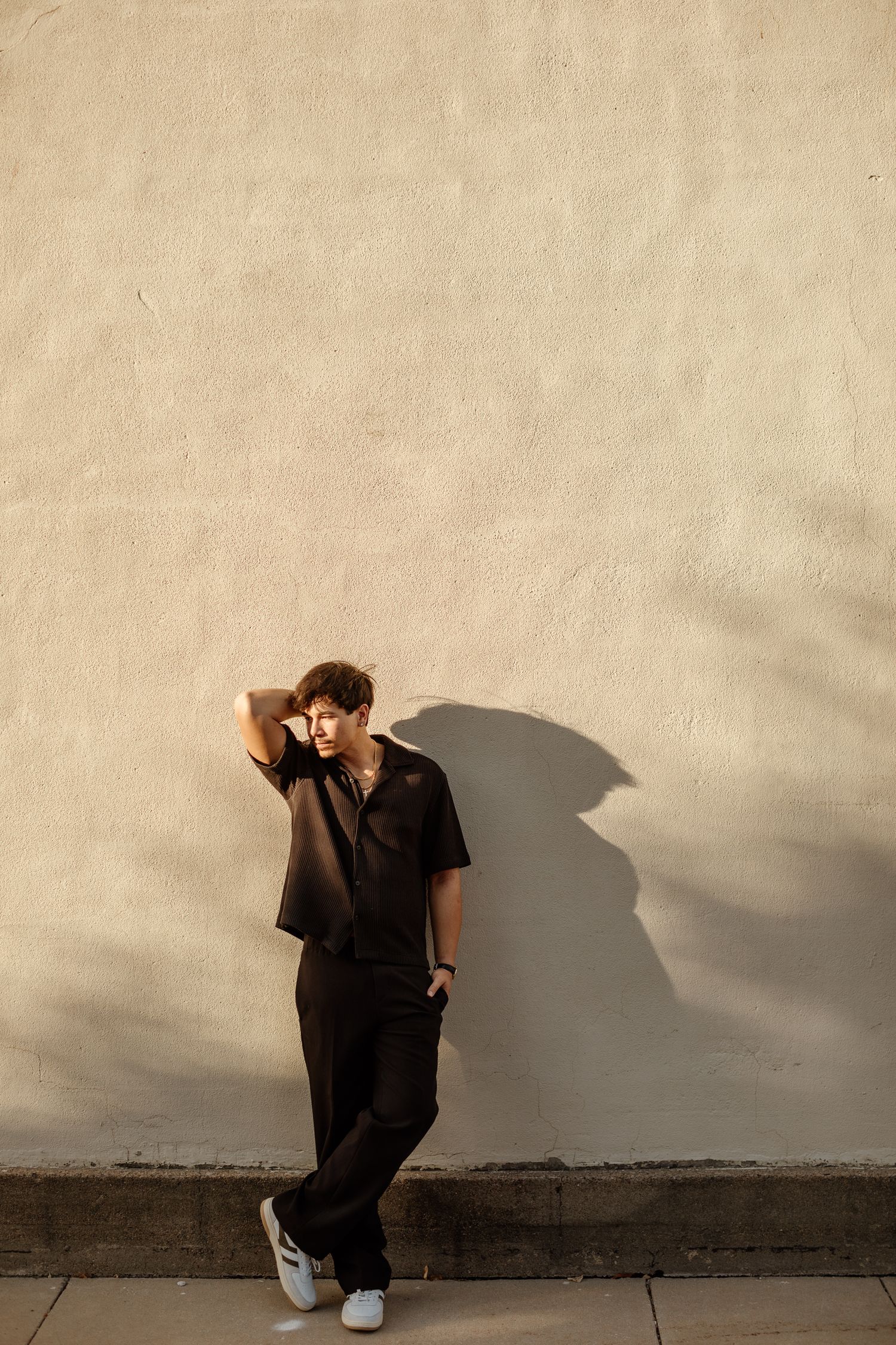 Stylish young man in all-black outfit leaning against beige stucco wall in golden hour sunlight.