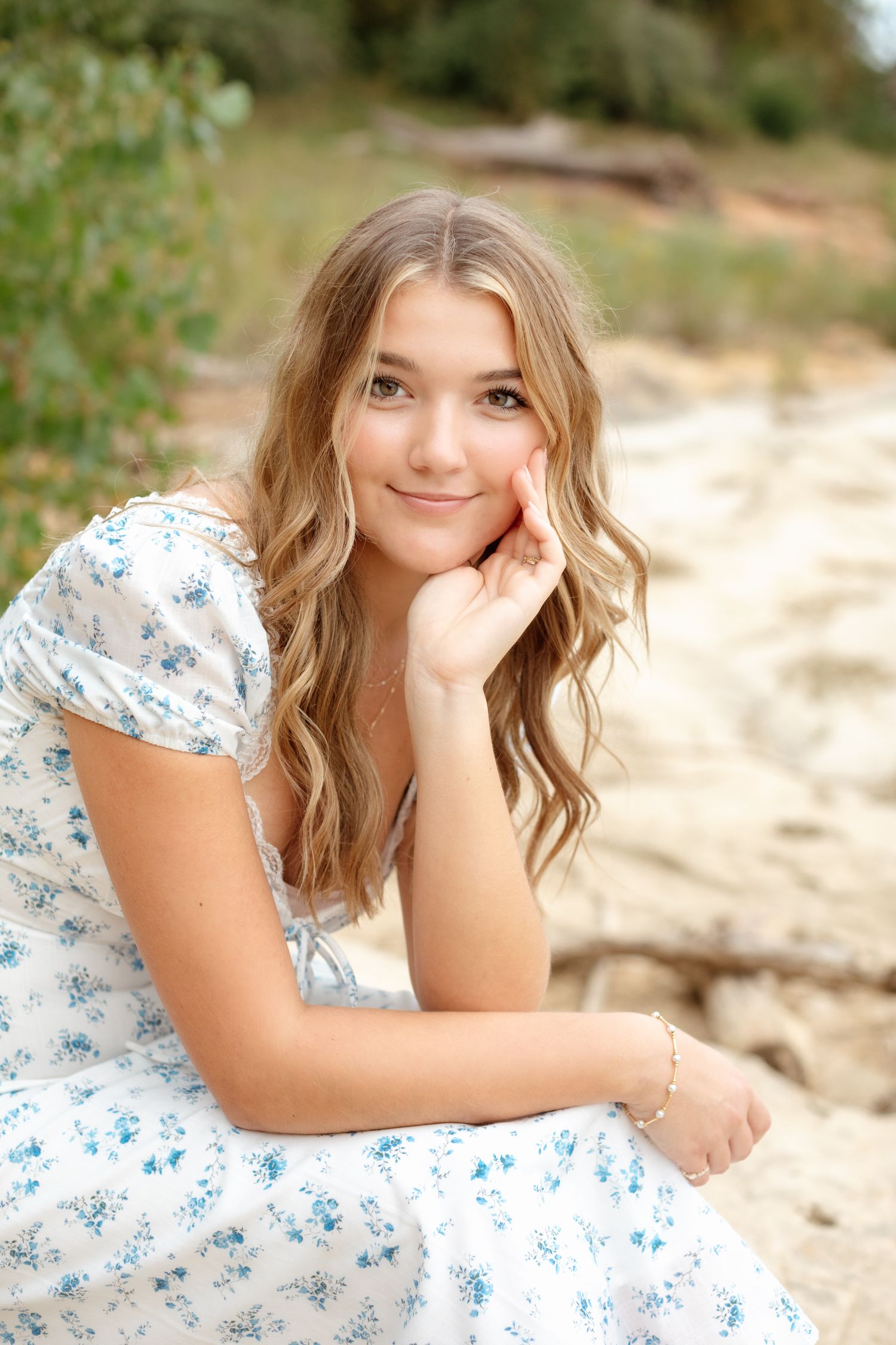 Smiling teenage girl in white floral dress resting chin on hand while sitting on sandy ground outdoors.