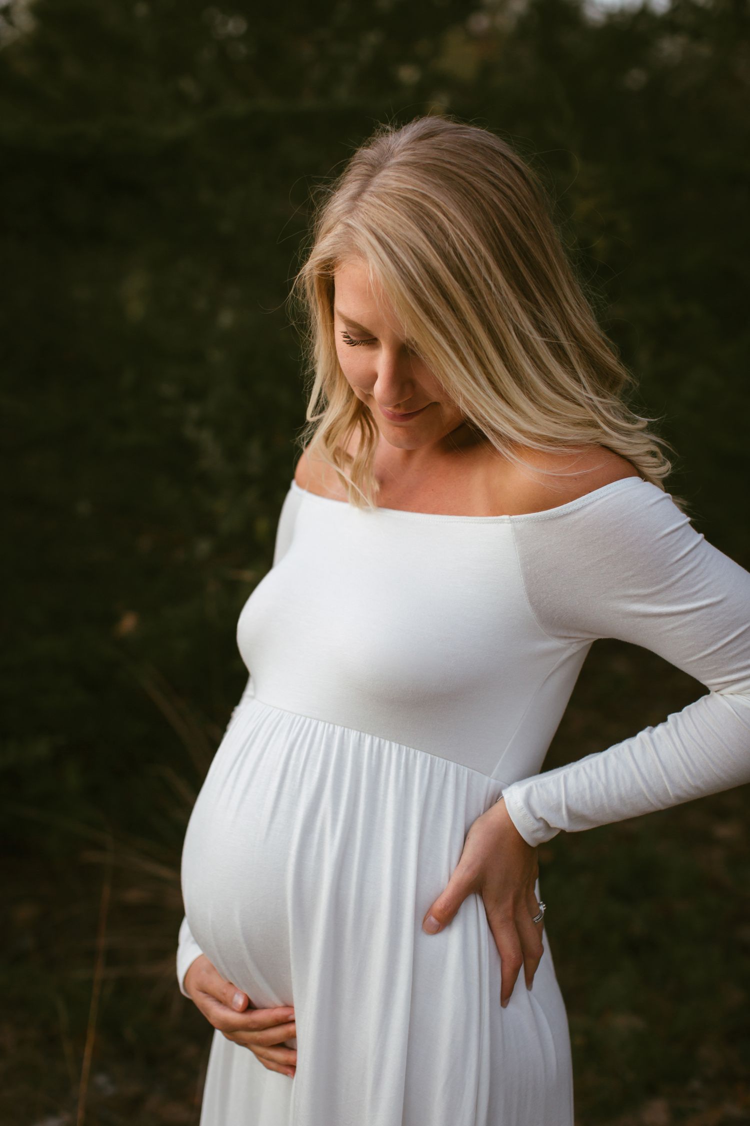 Pregnant woman in white off-shoulder maxi dress cradling baby bump outdoors with green foliage background.