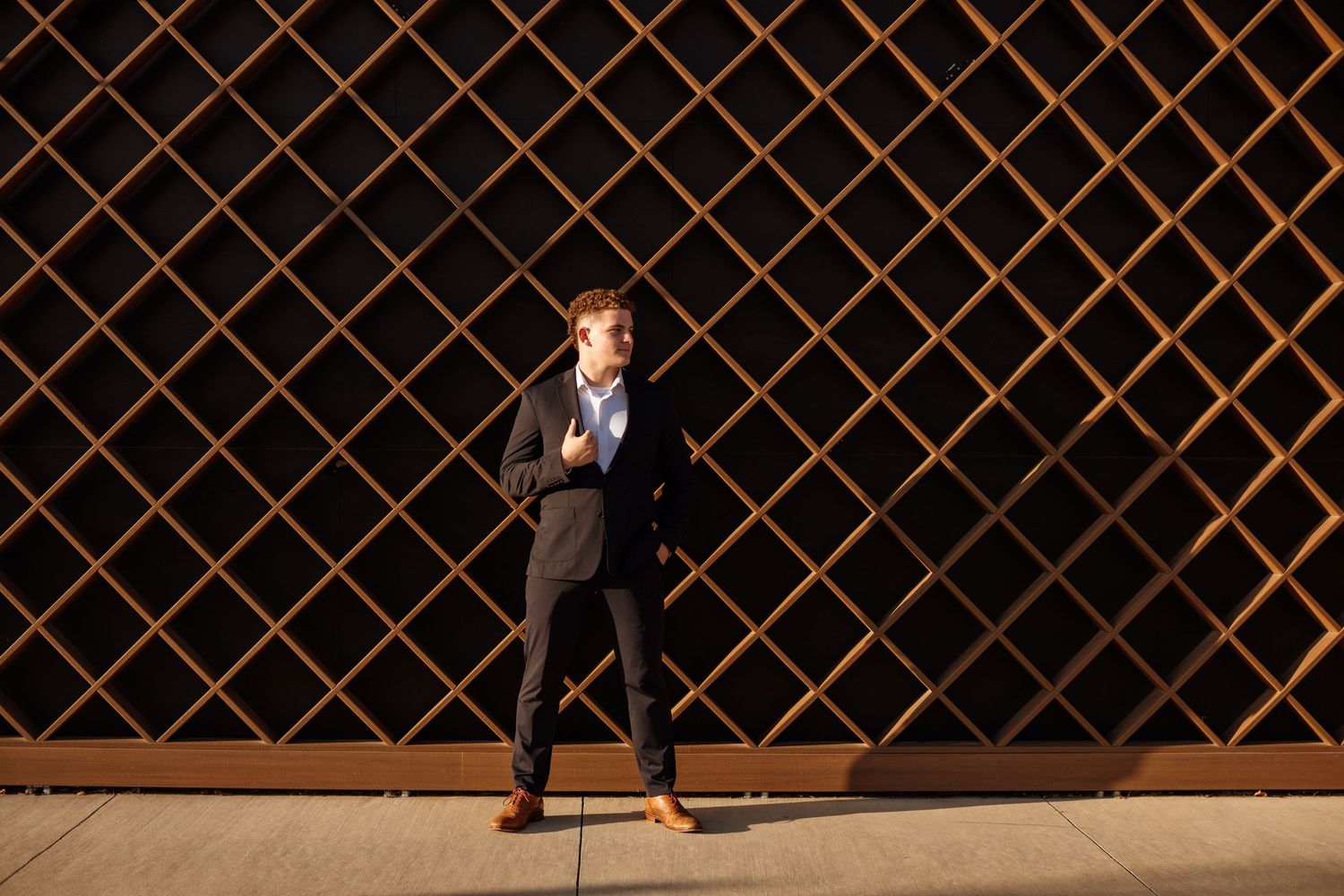 Confident young man in a dark suit posing in front of a geometric wooden lattice wall.