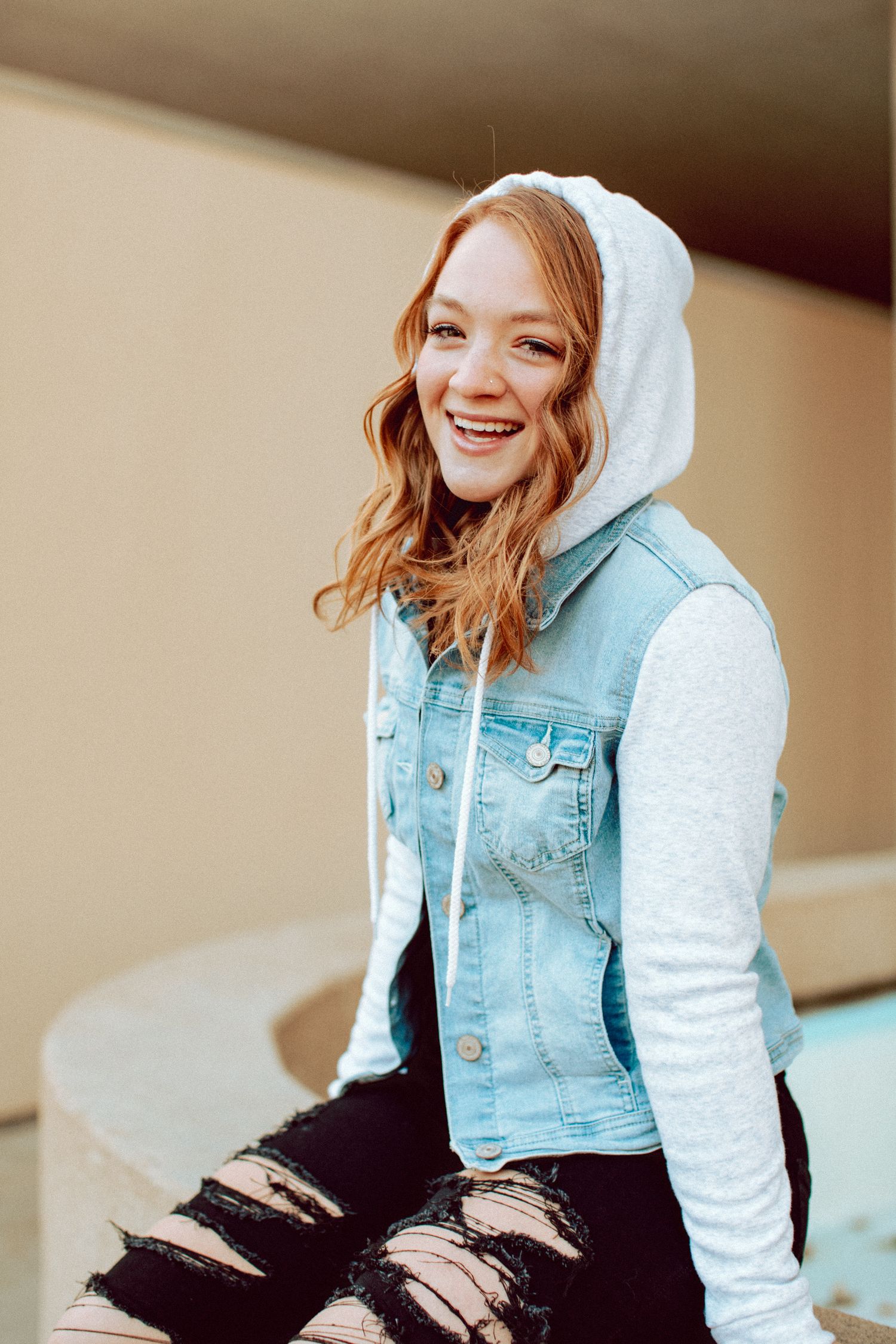Smiling teenage girl with red hair wearing a denim vest and gray hoodie sitting outside.