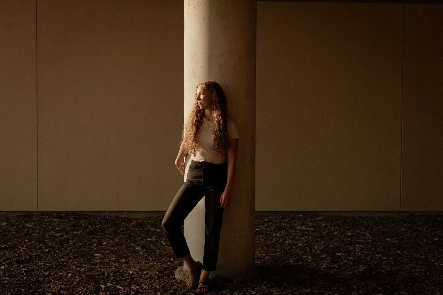 Young woman with long curly hair leans against a concrete pillar wearing white top and black pants.