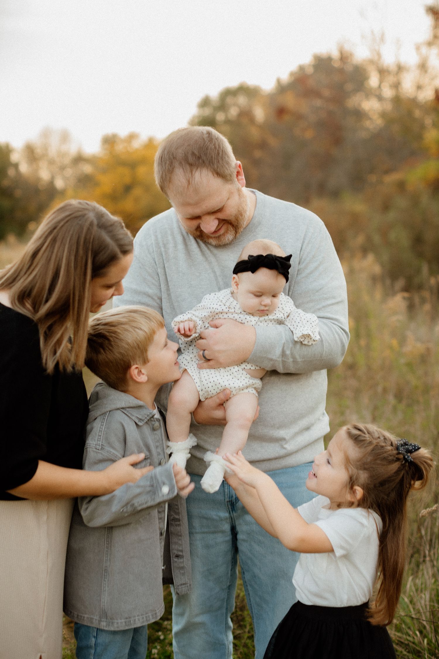 Joyful family of five gathered around father holding newborn baby outdoors in fall.