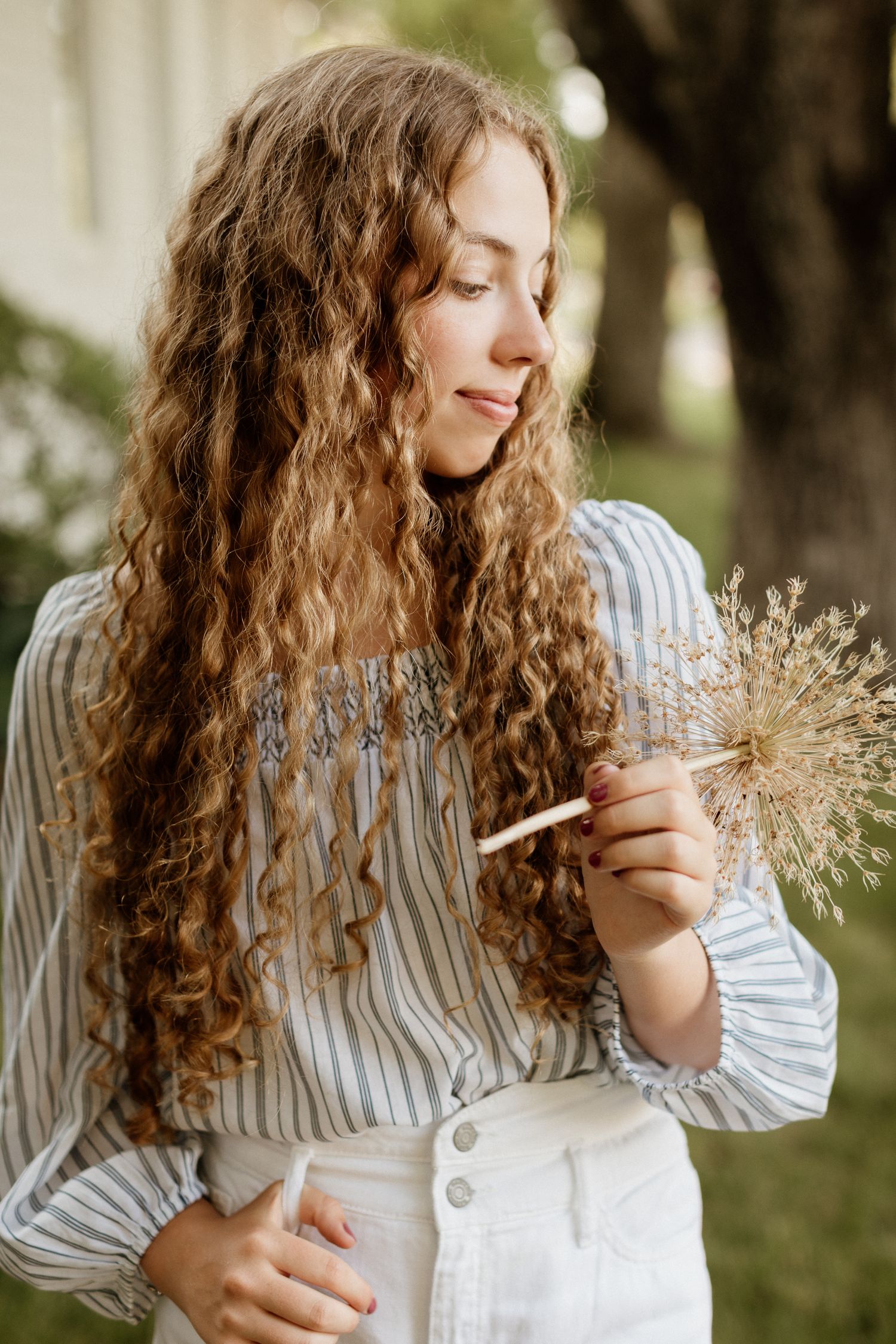 Young woman with long curly hair holds a dried allium flower, wearing a striped blouse outdoors.