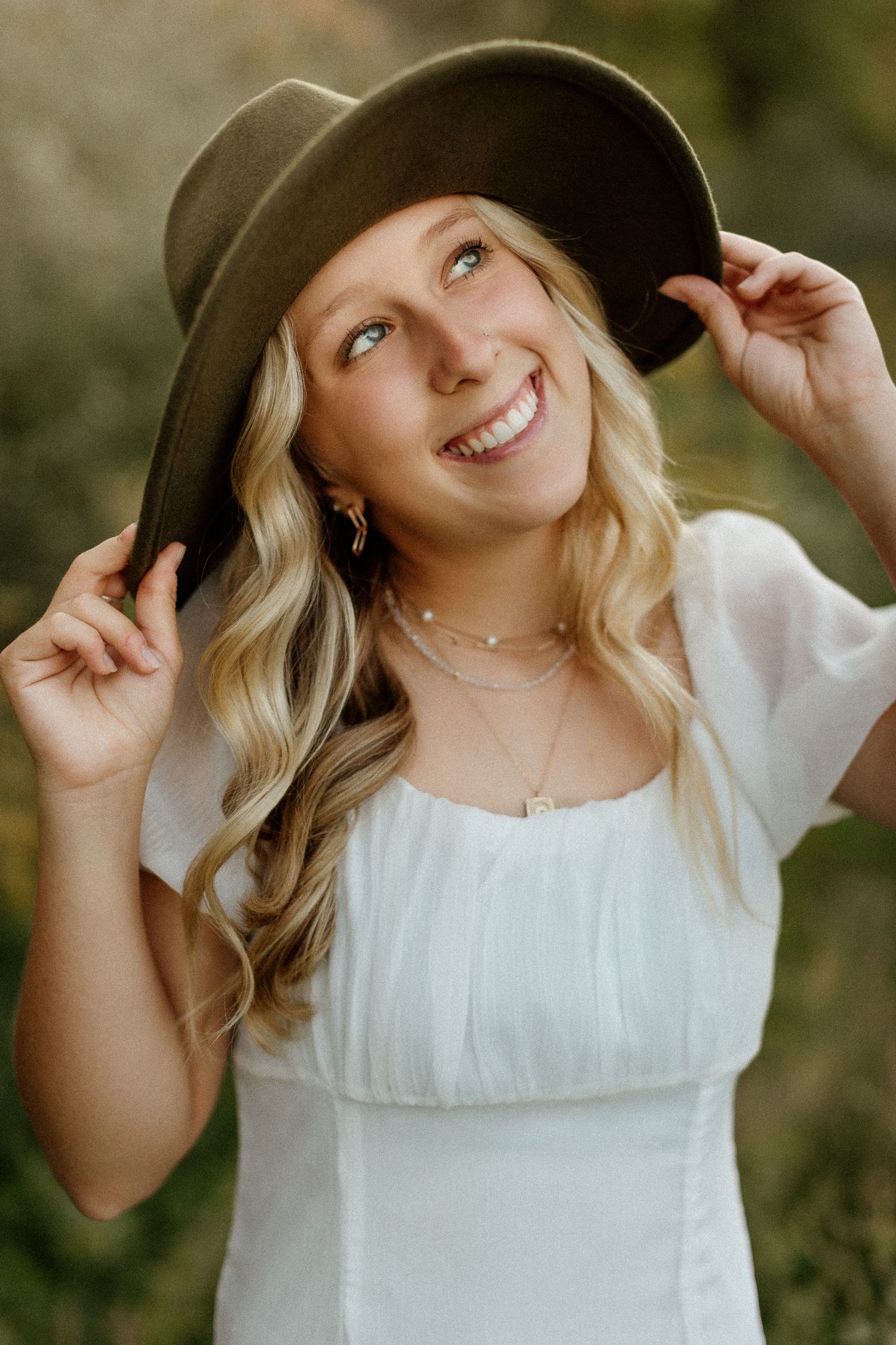 Blonde woman smiling joyfully while holding olive felt wide-brim hat in soft natural light.