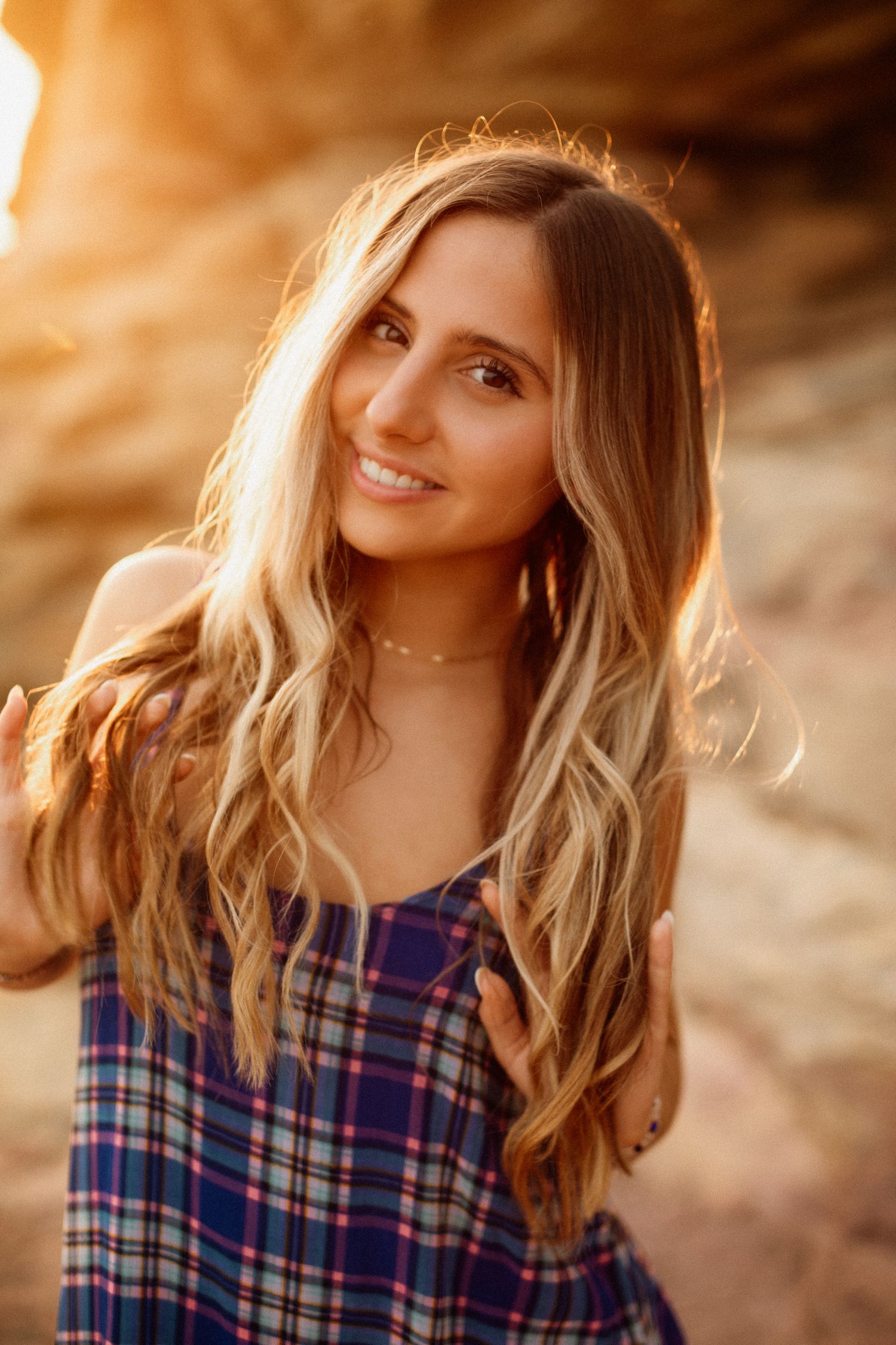 Young woman with long wavy blonde hair in plaid dress smiling in warm golden sunset light outdoors.