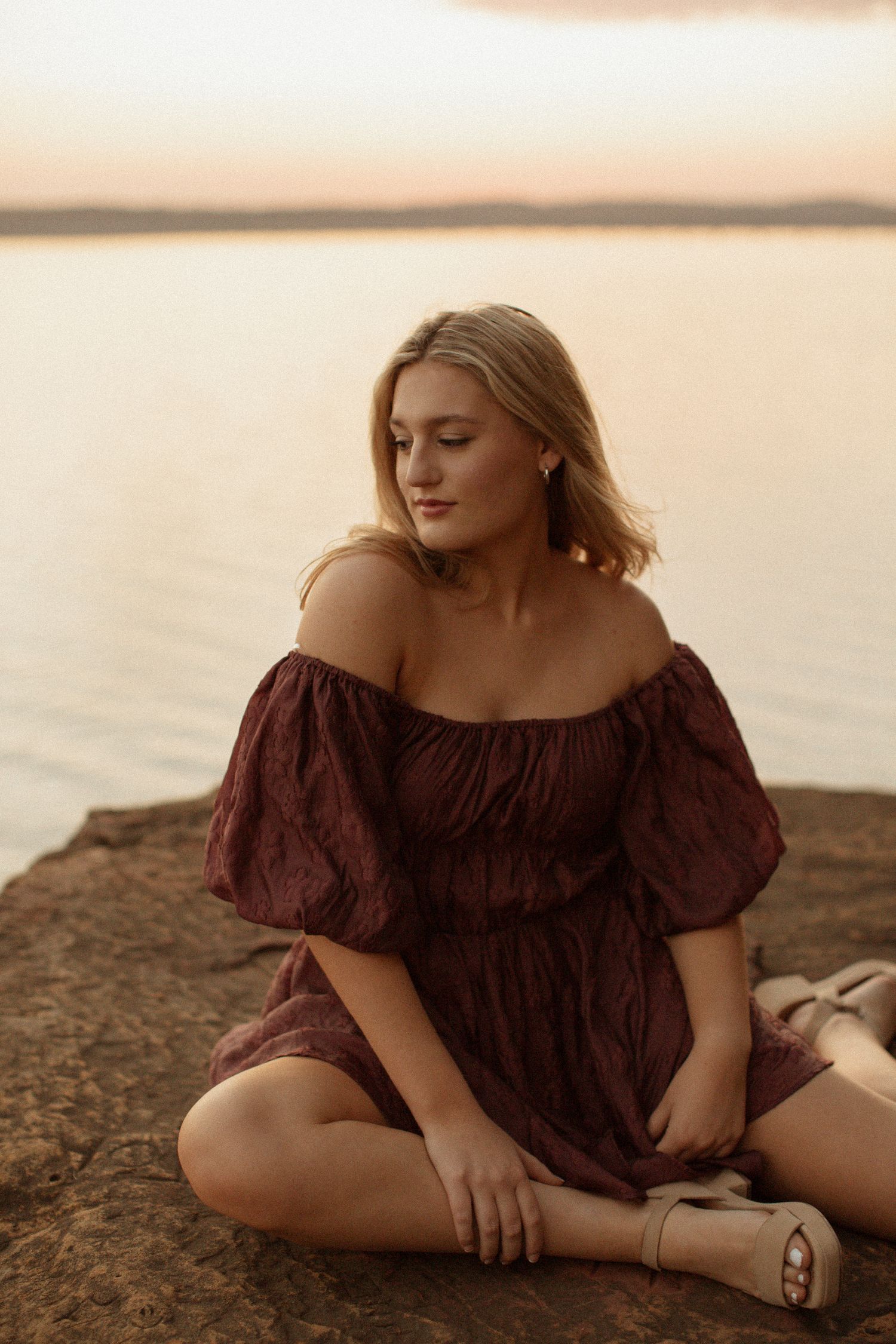 Blonde woman in a dark red off-shoulder dress sitting on rocks by a calm lake at sunset.