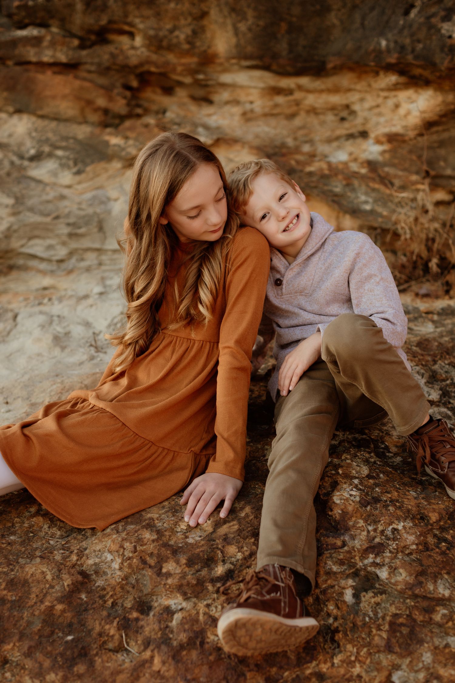 Young girl in an orange dress and boy in a beige sweater sitting together on rocky terrain.