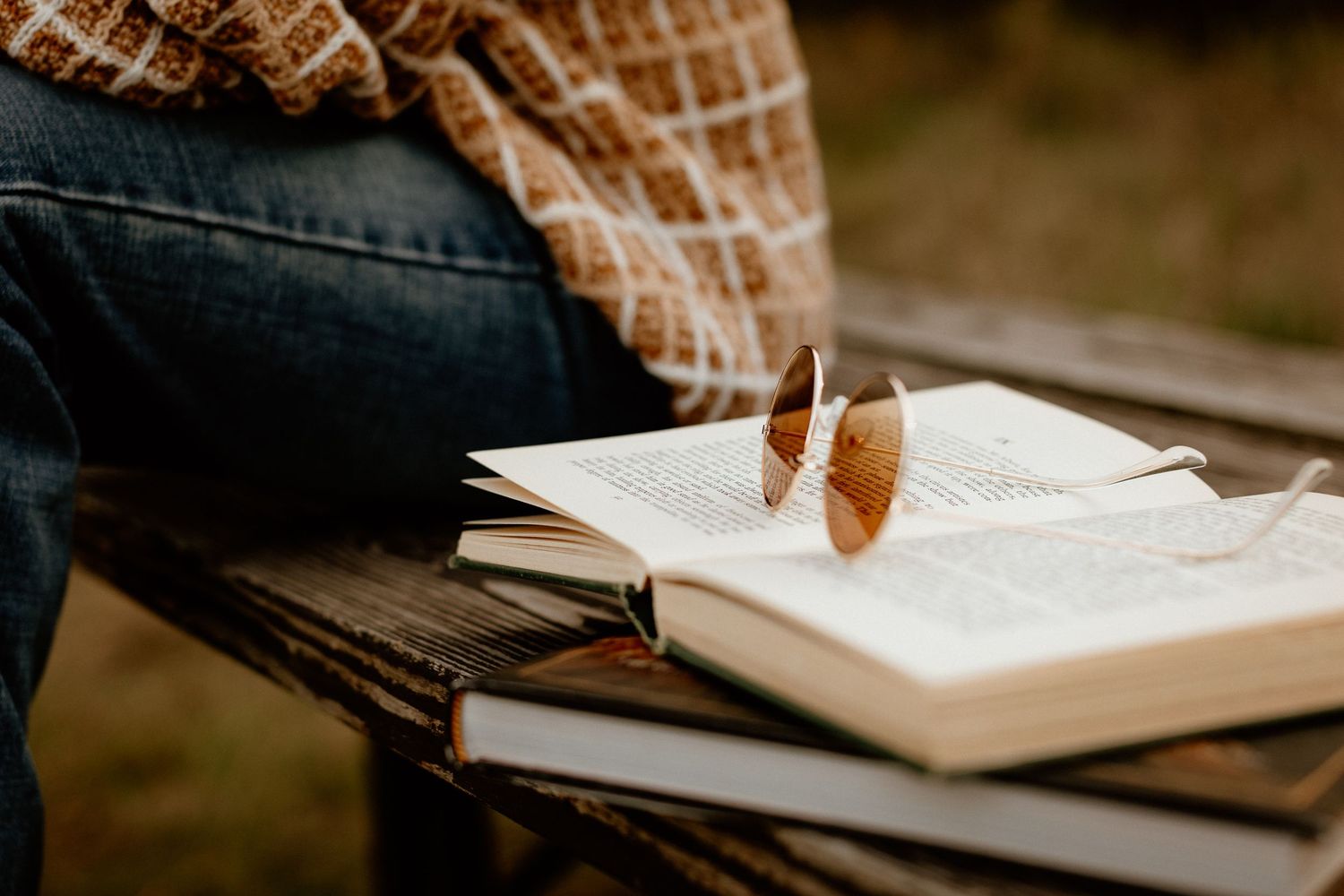 Open books with sunglasses resting on top, placed on a wooden bench outdoors in autumn.