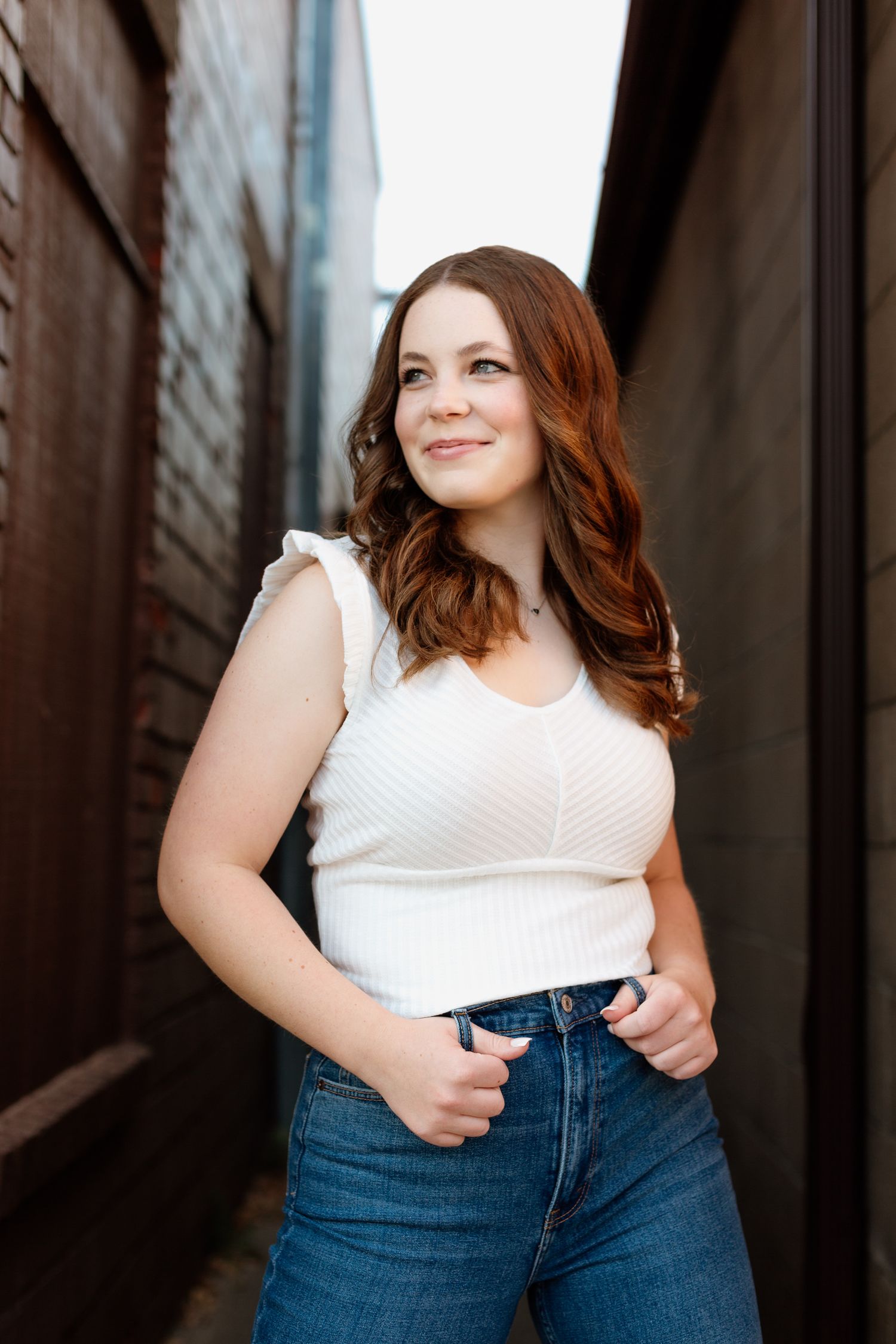 Young woman in white sleeveless top and jeans posing confidently in narrow brick alleyway.