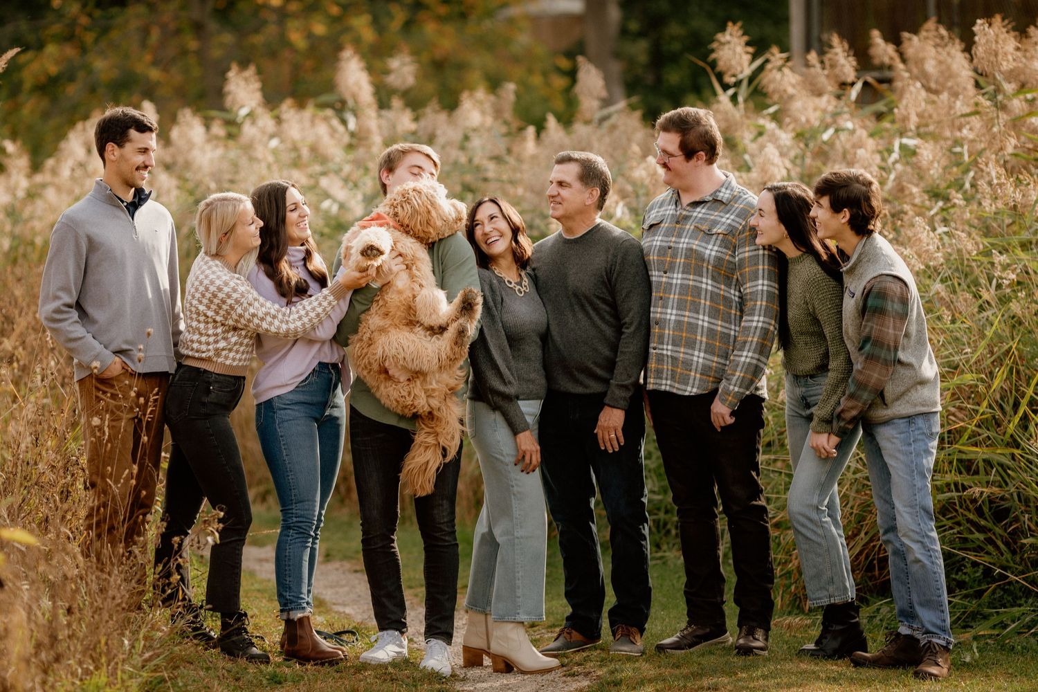 Large extended family group smiling together outdoors with a dog in fall foliage.