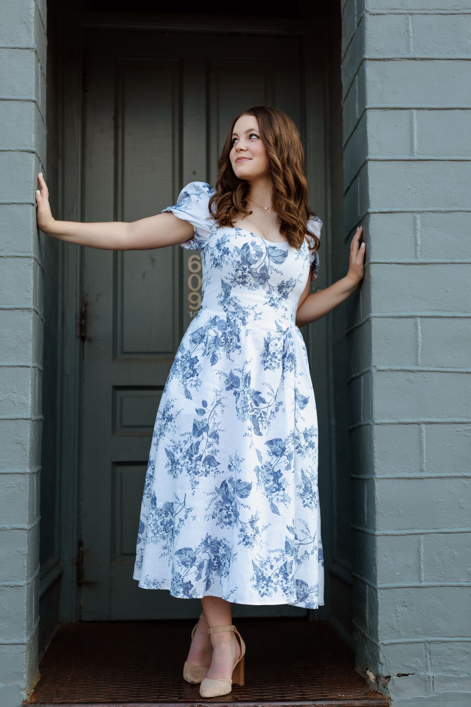 Young woman in white blue floral midi dress posing in doorway of gray brick building.