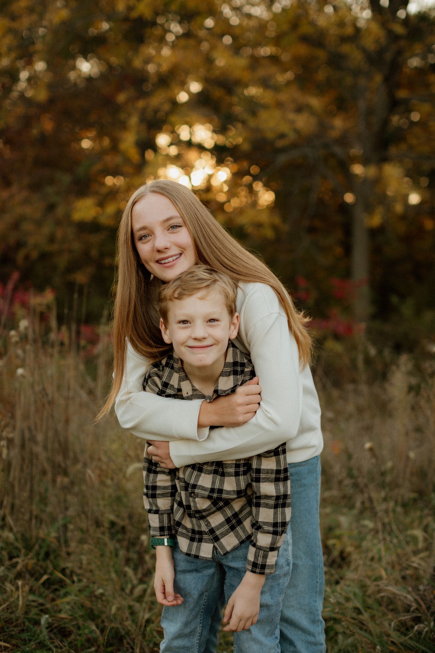 A teenage girl hugs a young boy from behind, both smiling outdoors in a fall setting with golden foliage.