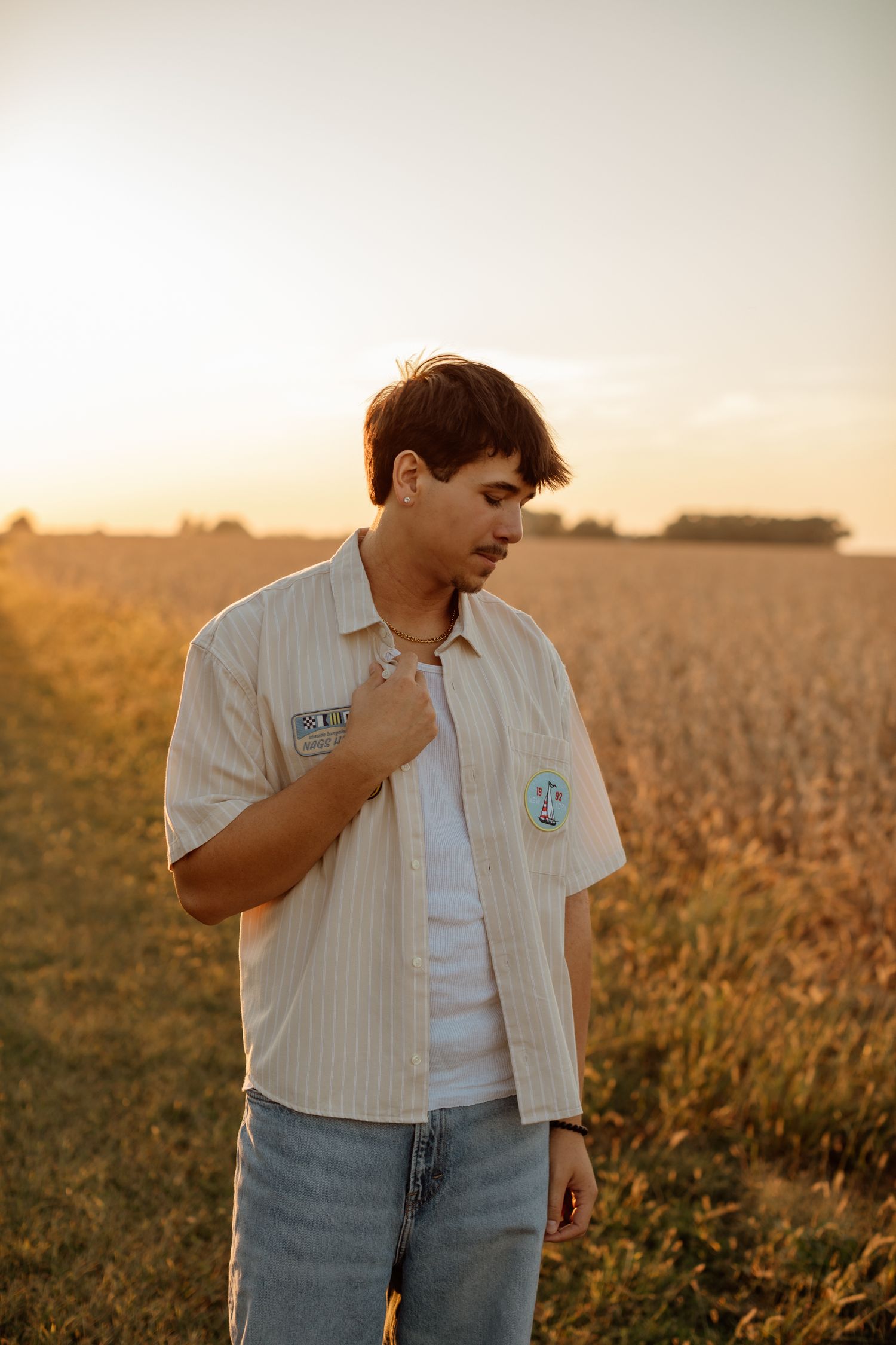 Young man in striped button-up shirt standing in golden sunset light overlooking an open farm field.