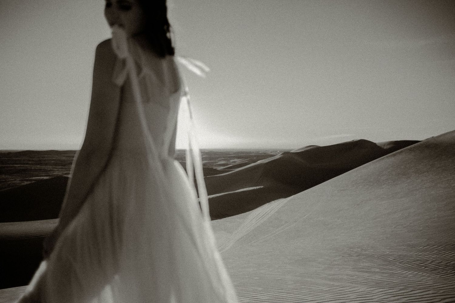 Woman in a flowing white dress standing among sand dunes in an artistic black and white photo.