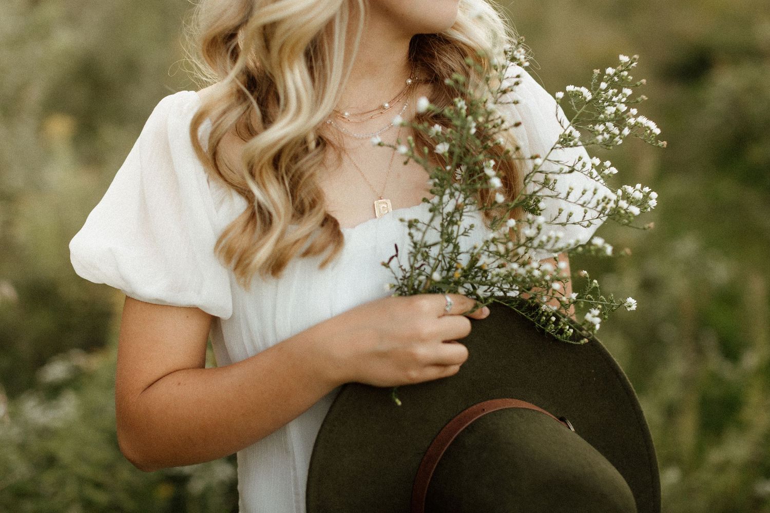 Blonde woman in white dress holding wildflowers and a dark hat in a field.