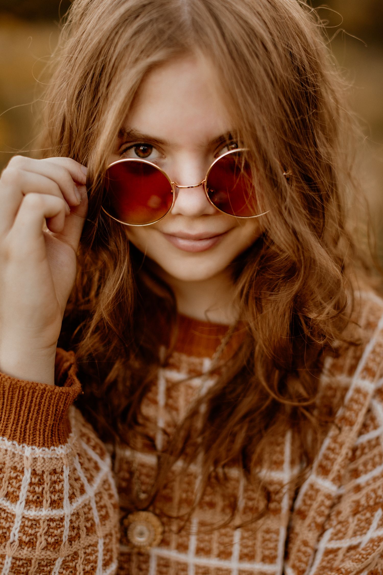 Young woman with wavy brown hair peers over tinted round sunglasses, wearing a cozy brown patterned sweater.