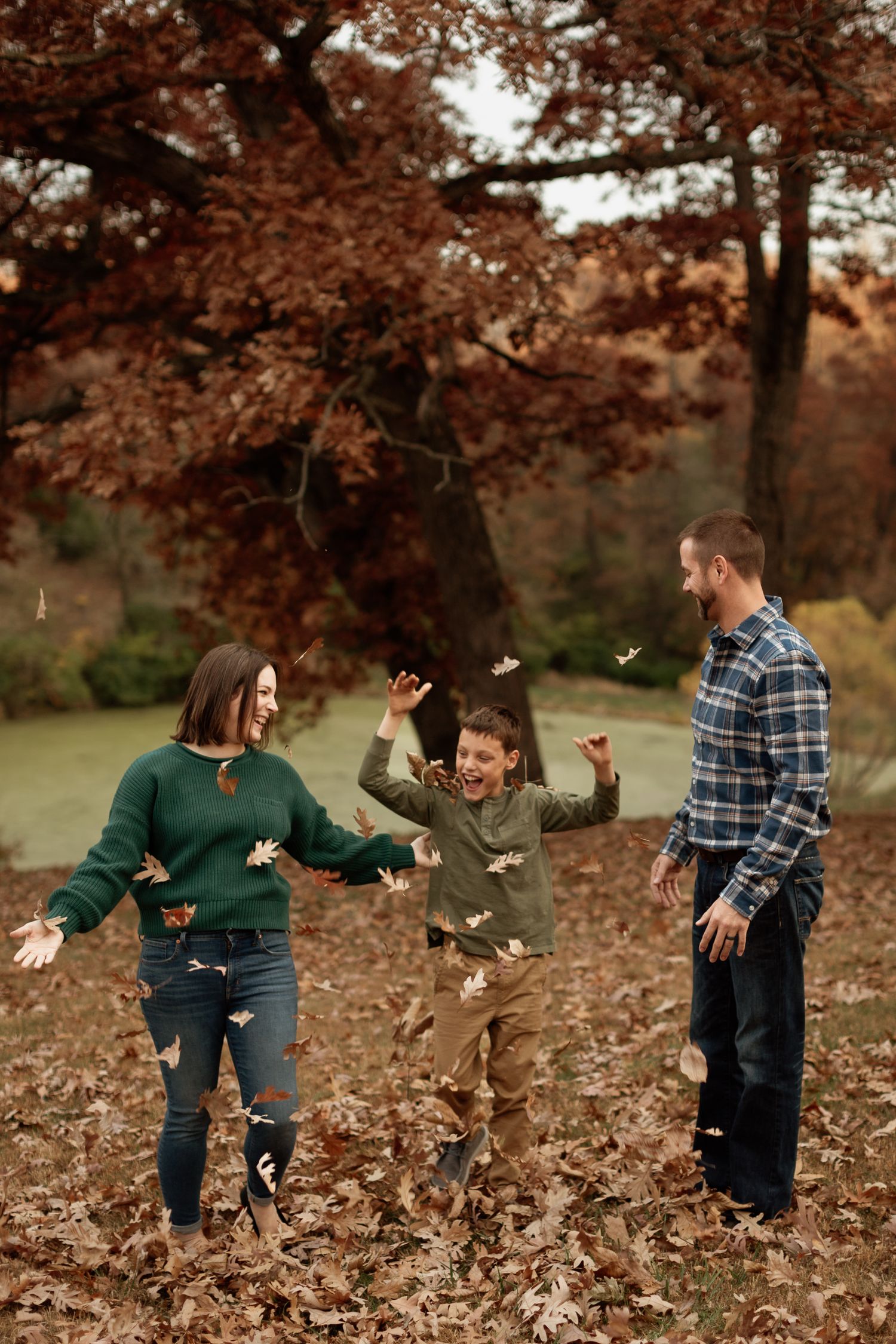 Joyful family of three tossing autumn leaves in the air under a red tree.