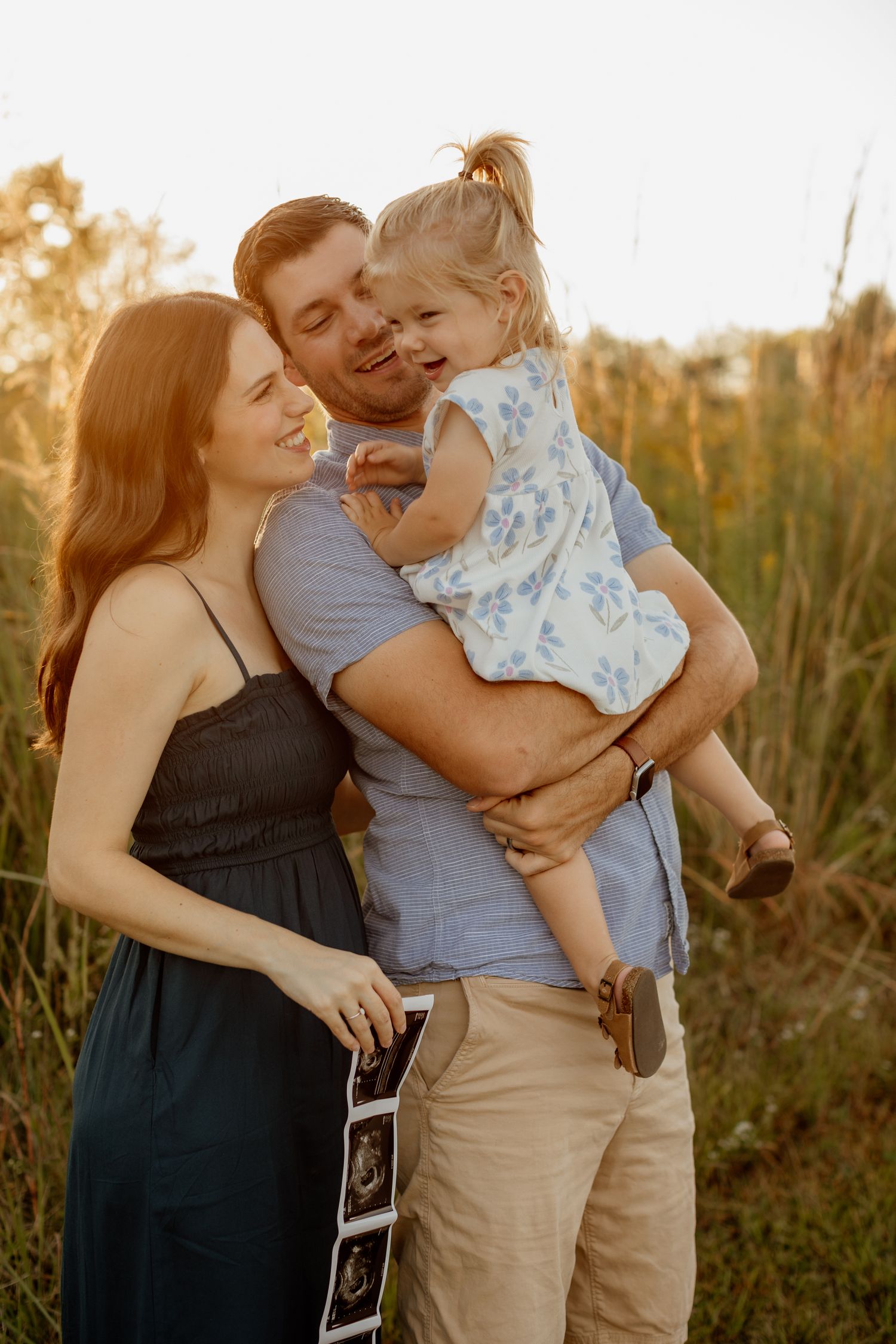Happy family with toddler holding ultrasound photos in a golden sunlit field.