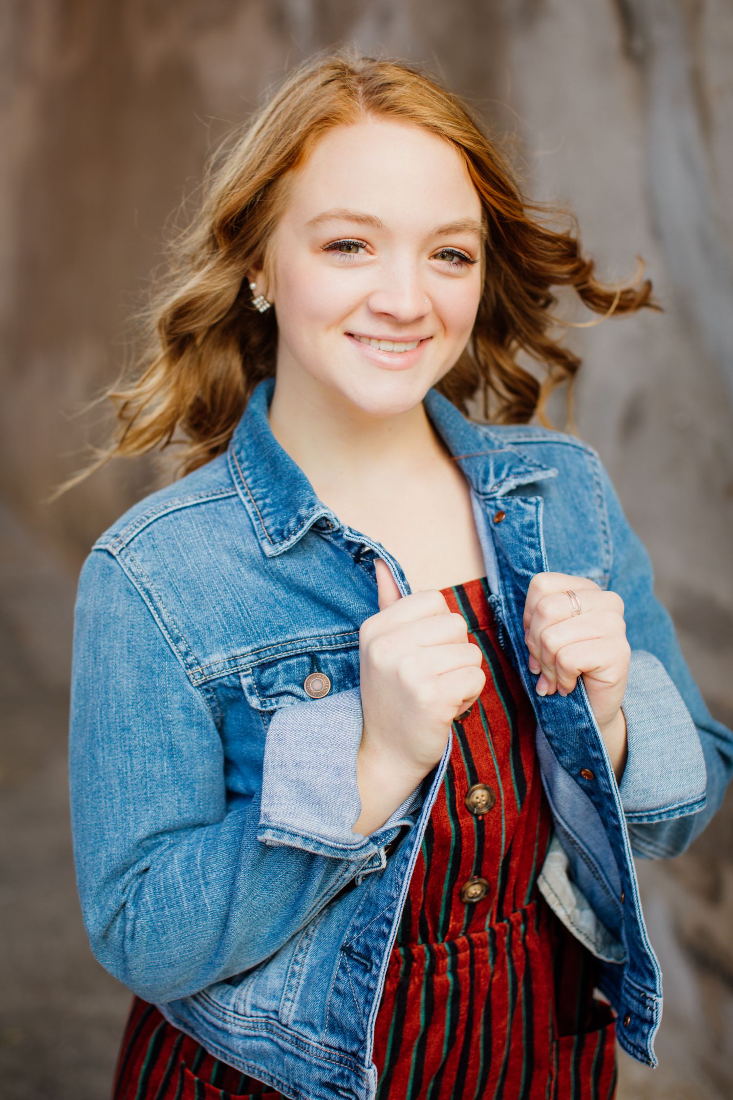 Smiling redhead teen in denim jacket over striped jumpsuit poses confidently outdoors.