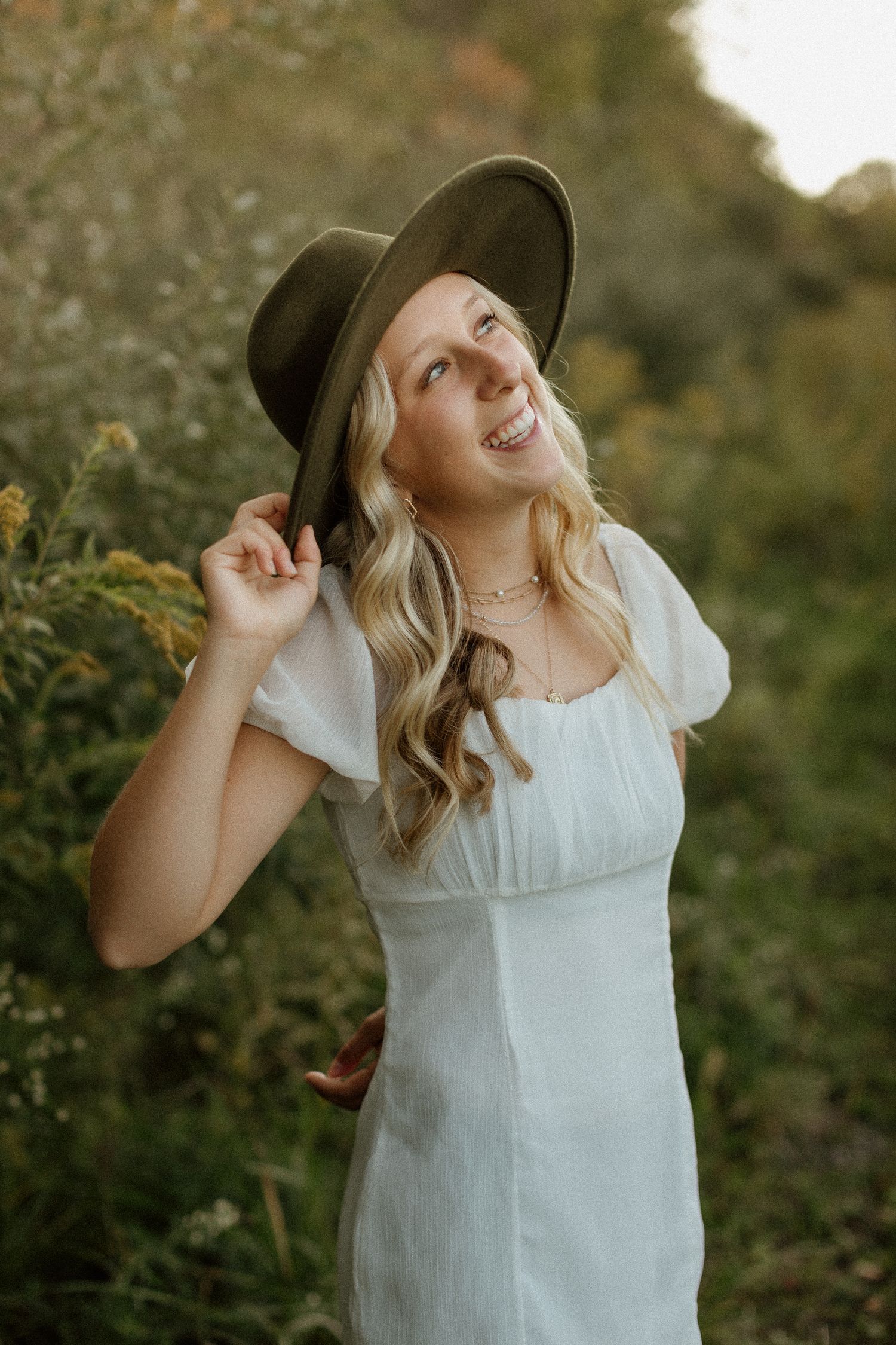 Smiling blonde woman in white dress holding olive wide-brim hat outdoors in golden light.