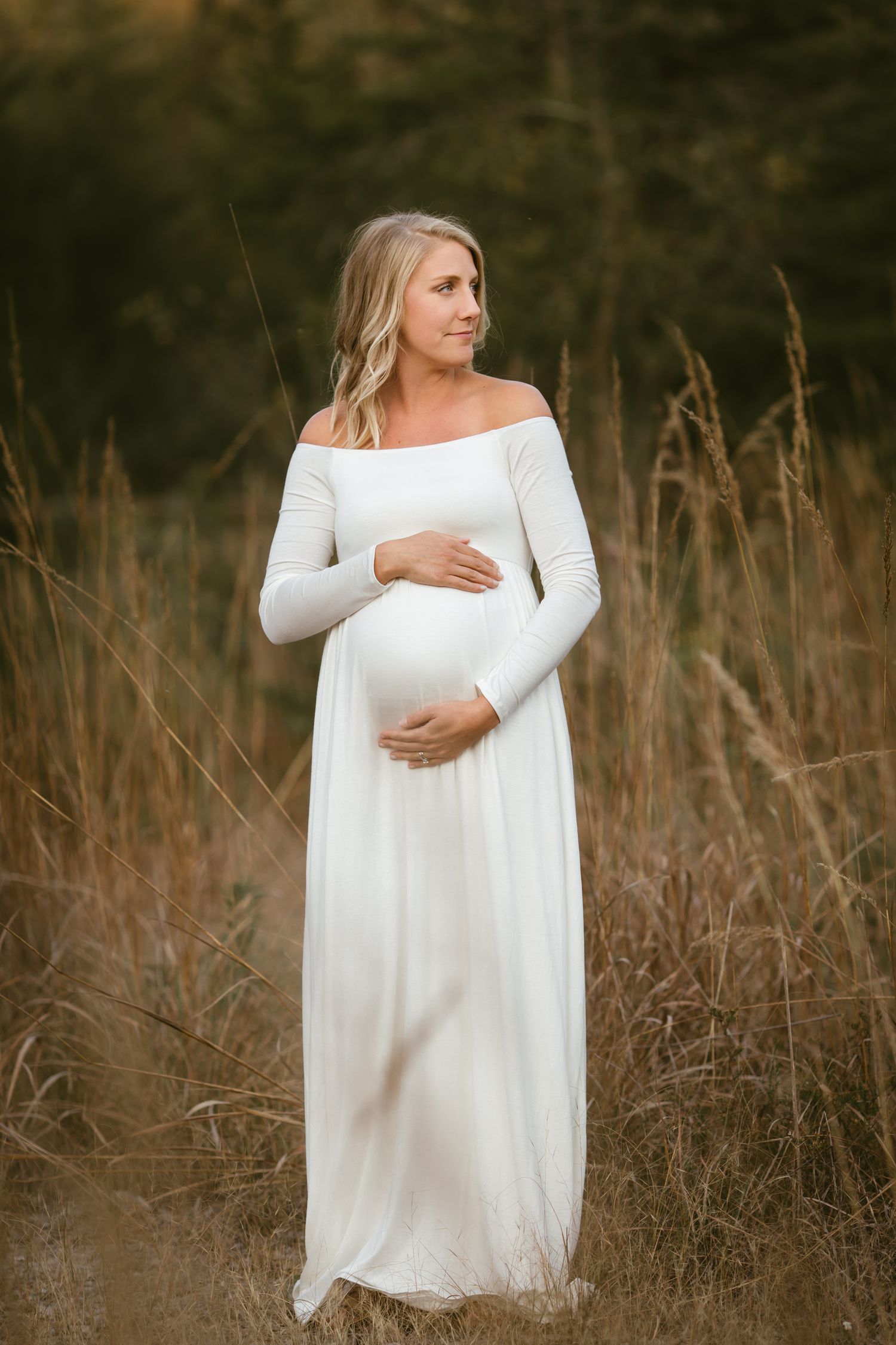 Pregnant woman in white off-shoulder gown cradling baby bump in golden field at sunset.