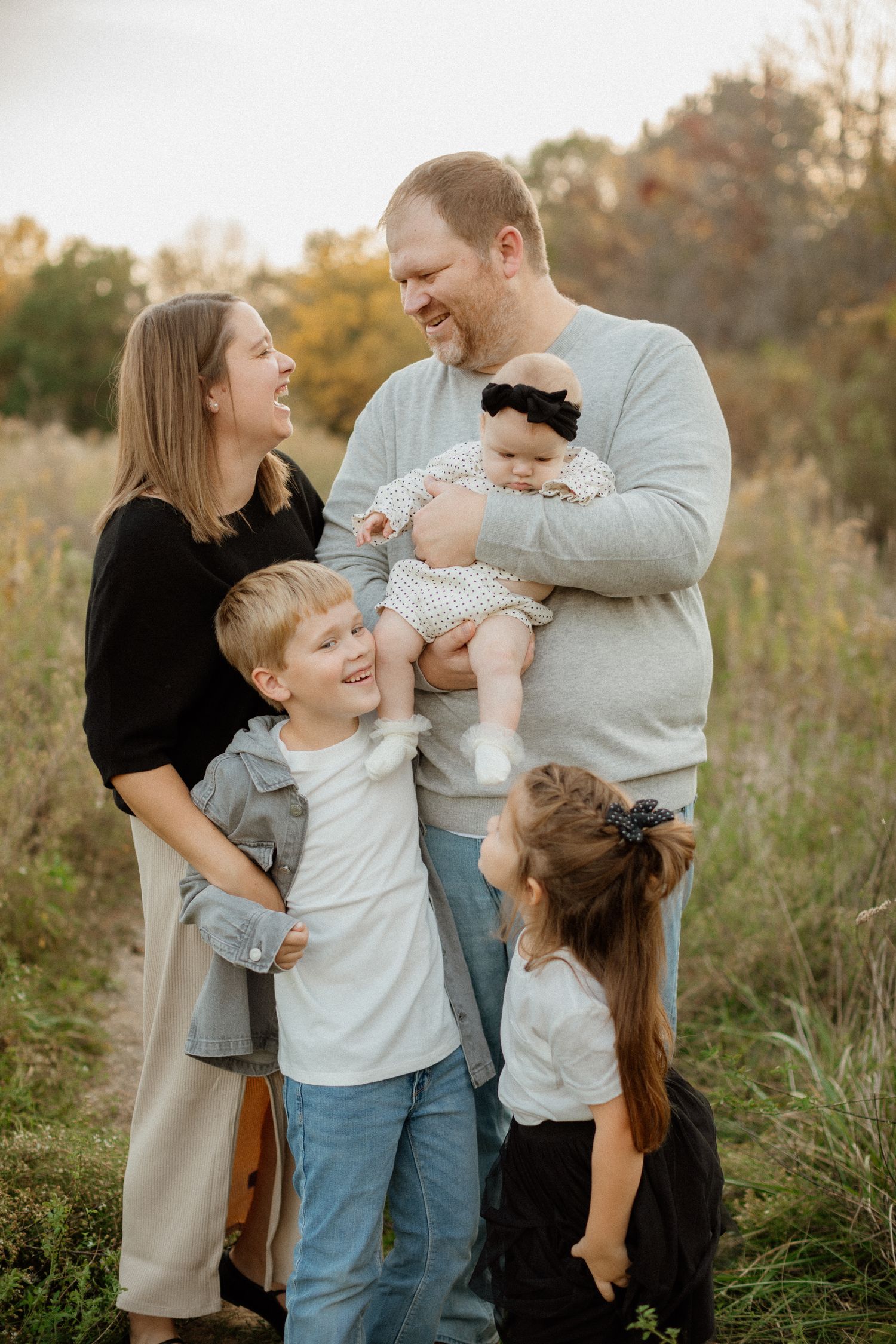 Laughing family of five with newborn baby posing together in an autumn field.