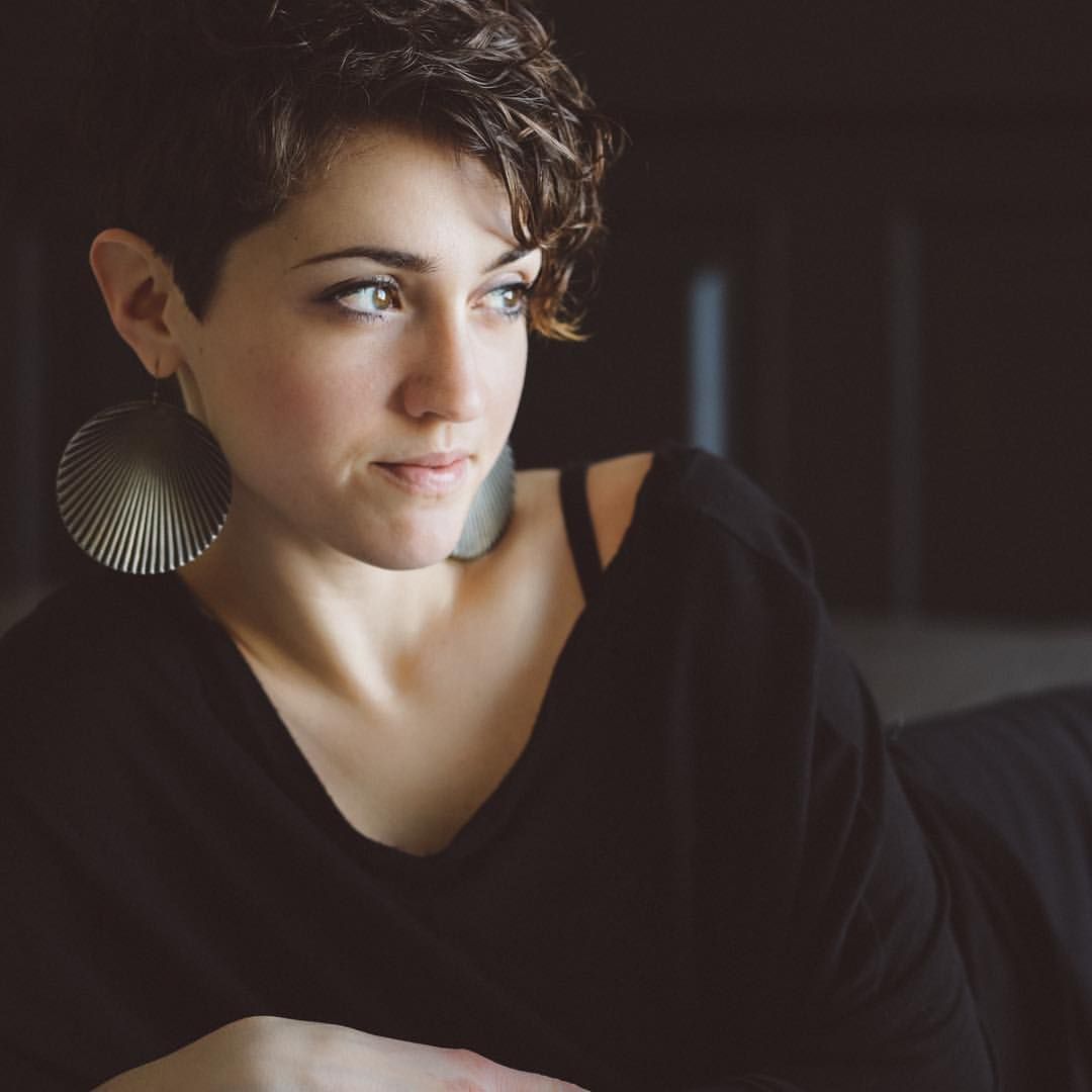 Young woman with short curly hair and large earrings gazing thoughtfully in soft light.