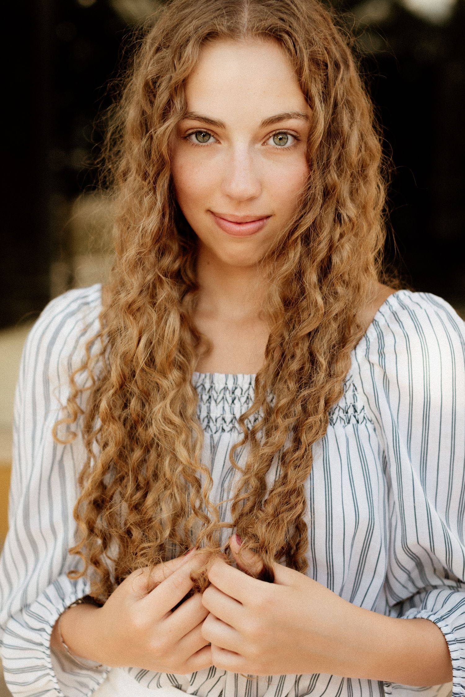 Young woman with long curly blonde hair wearing a striped blouse in a close-up portrait.