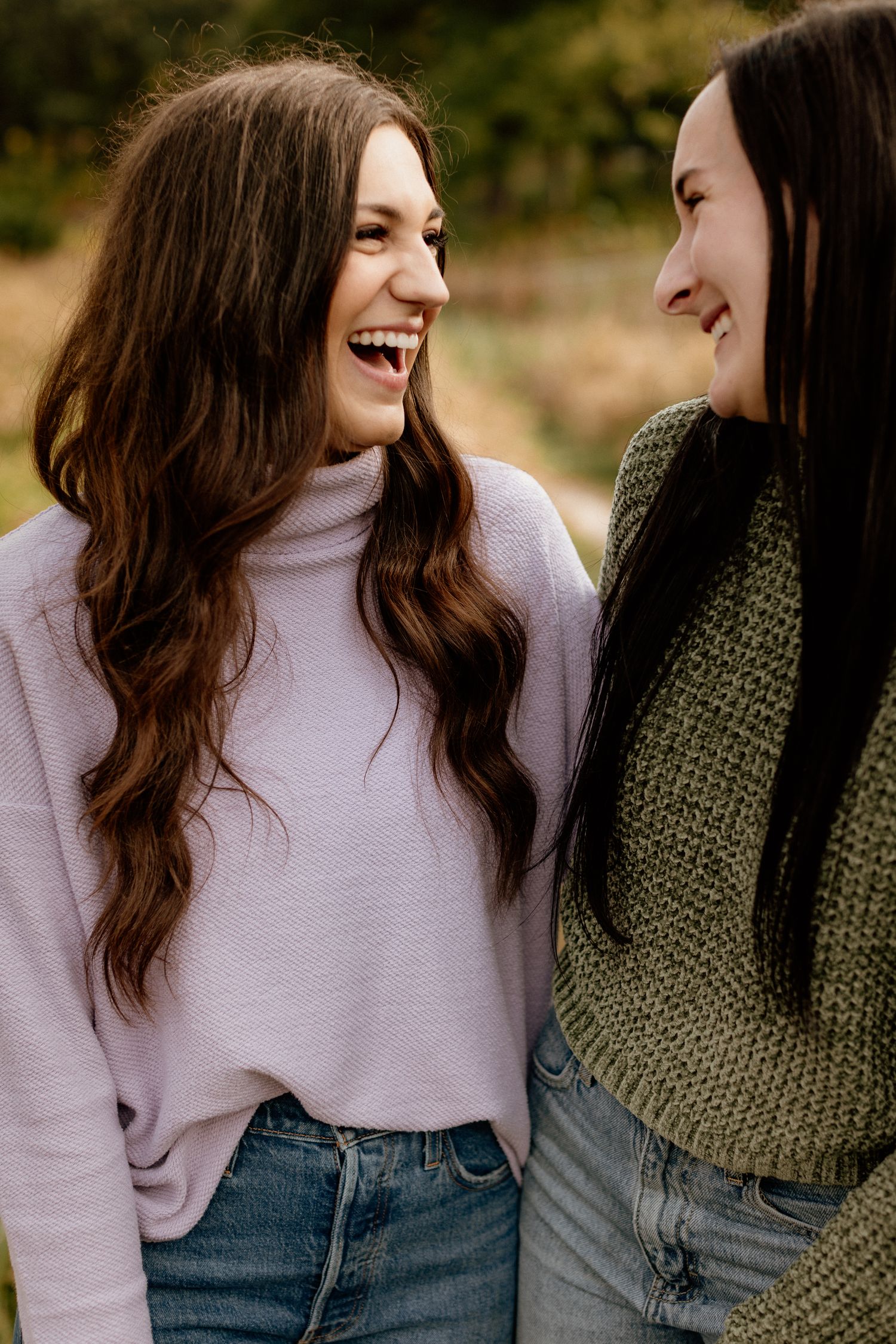 Two young women laughing and smiling at each other outdoors in a fall setting.