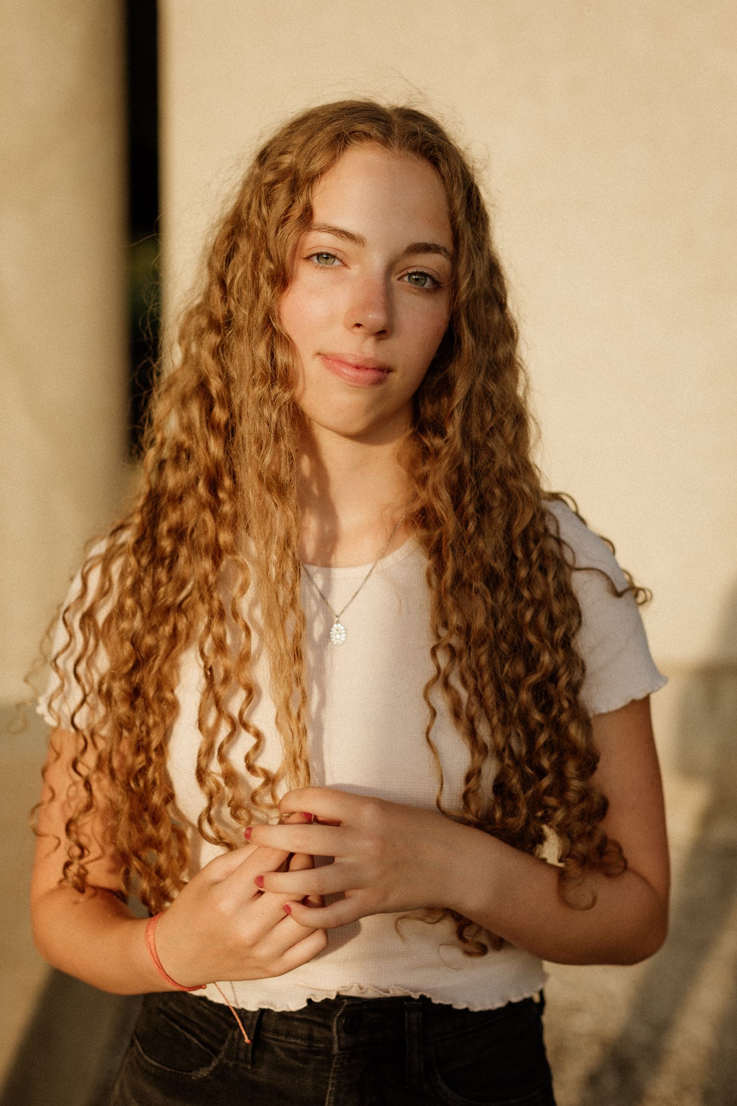 Young woman with long curly auburn hair poses in a white t-shirt and dark jeans in warm sunlight.