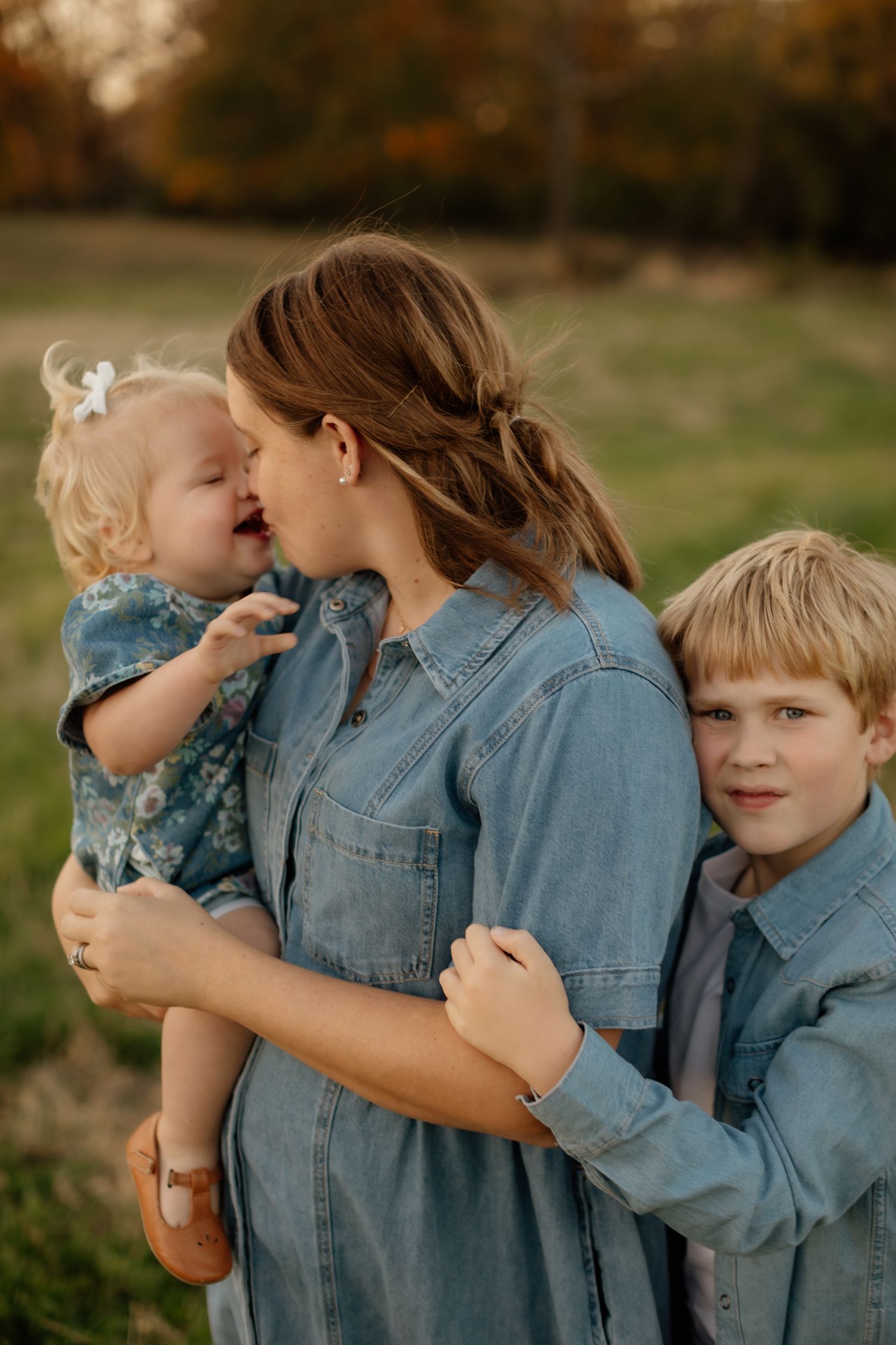 Mother in denim dress holding toddler while older child embraces her in a field.